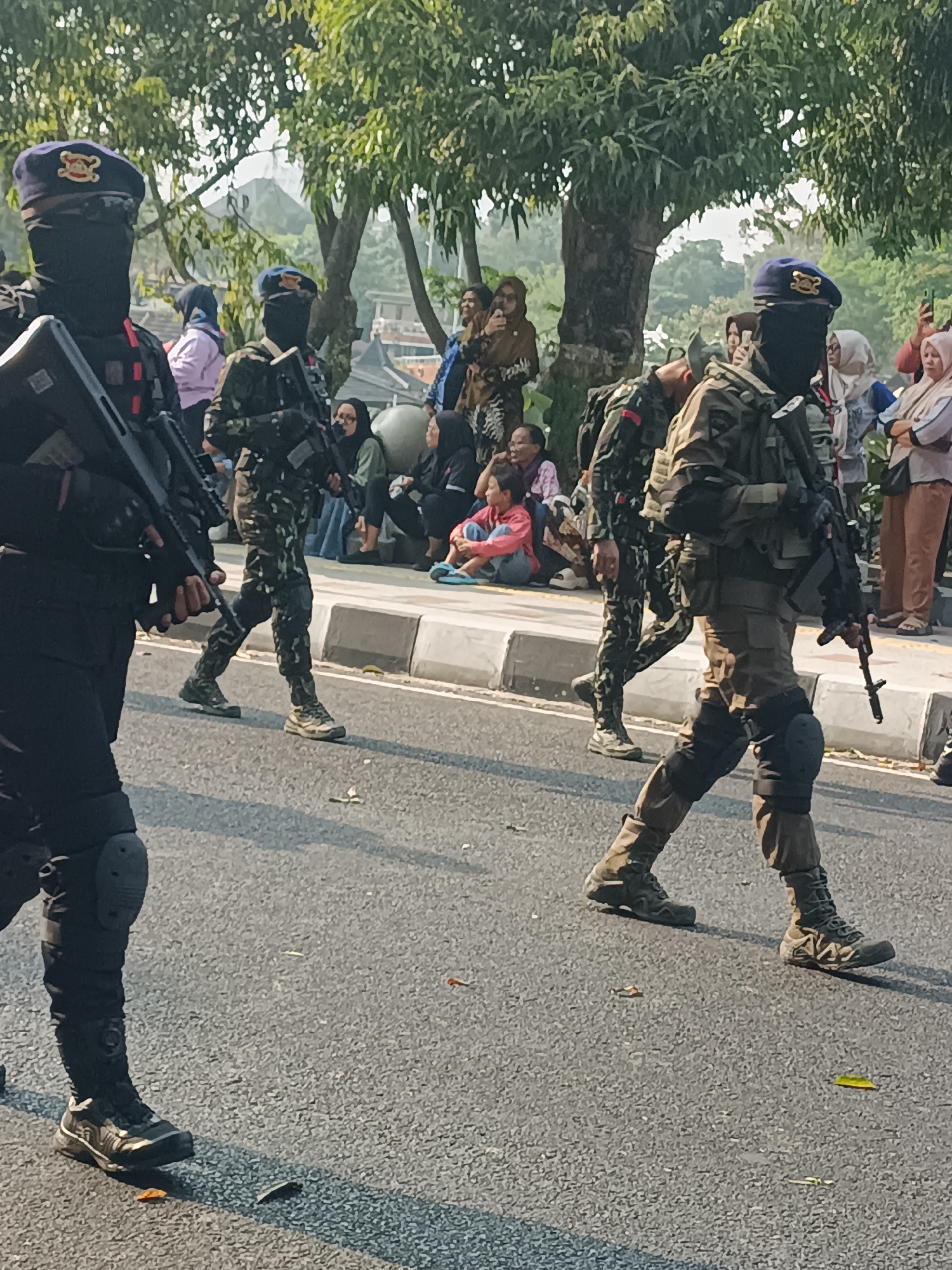 This image captures a street scene in Sleman, Indonesia, where individuals in tactical gear, armed with rifles, are escorting or patrolling. Two prominent figures in the foreground, wearing black masks that obscure their faces, blue berets with insignia, and camouflage uniforms, are walking on a paved road. The individual on the left carries a rifle with a black stock and magazine. The person on the right also carries a rifle, with a tan and black color scheme. Both are equipped with tactical vests, knee pads, and sturdy footwear.
Behind them, a crowd of civilians is gathered on a raised sidewalk. Among them are women and children, some observing the scene, and others appearing to engage in conversations. The environment suggests a public space with trees and foliage providing shade, and buildings in the background, indicating an urban or semi-urban setting. The sunlight casts shadows, indicating it is daytime. The presence of the armed individuals suggests a security operation or public event requiring a visible security presence in Sleman.