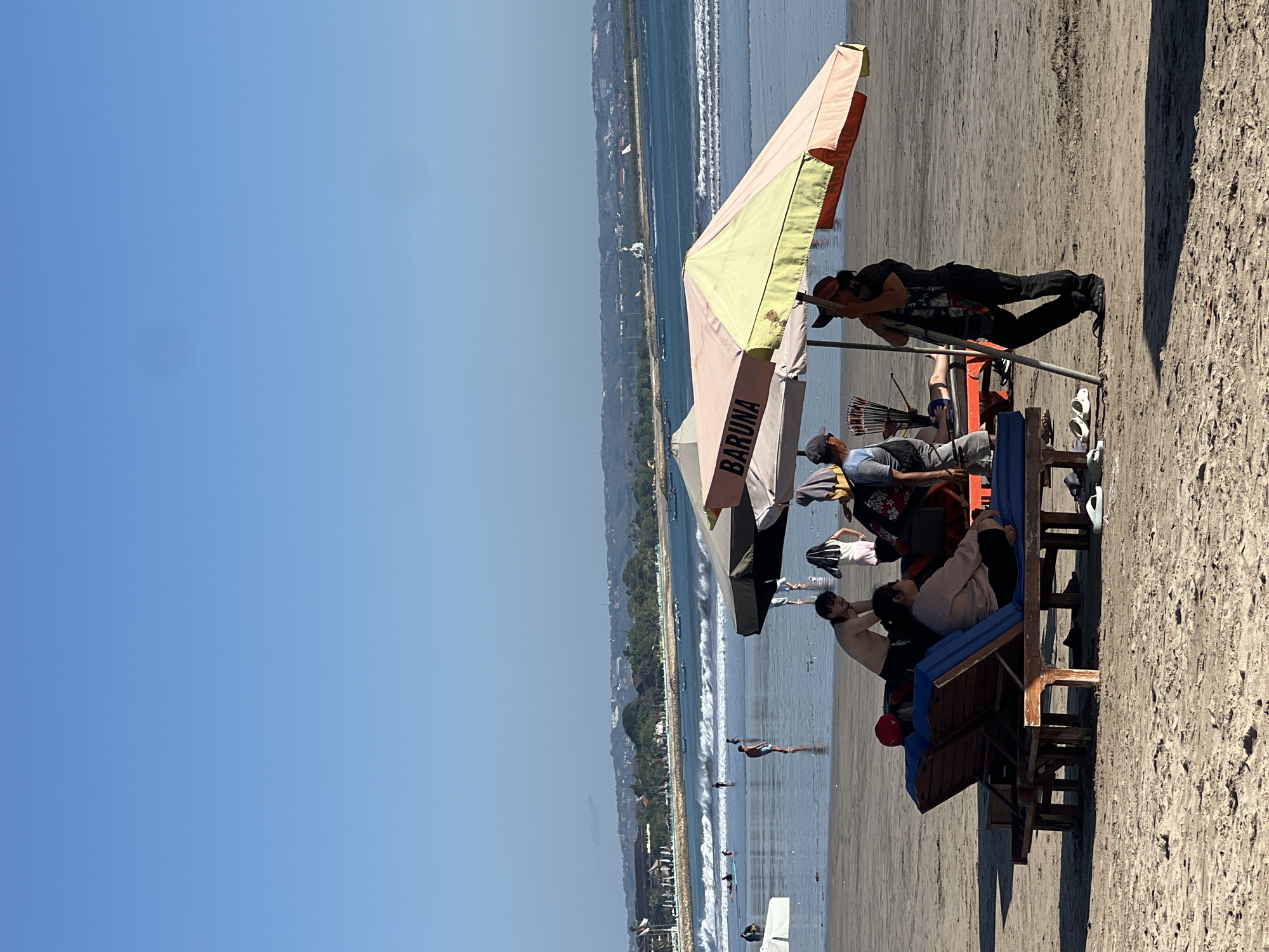 The image depicts a beach scene in Kuta, Indonesia, under a clear blue sky. In the foreground, two wooden lounge chairs are occupied by individuals. One chair has a person wearing a red cap and a pink top, while the other has a person in a pink sweatshirt and black pants. Adjacent to the chairs, a vendor stands under a multi-colored umbrella that reads "BARUNA" on the side. The vendor is dressed in black pants and a dark top, holding a bundle of items. Other people are visible in the midground, some standing near the water and others walking along the shoreline. The ocean in the background features moderate waves, and the coastline is lined with trees and buildings. The sand on the beach shows footprints and the texture of the grains.