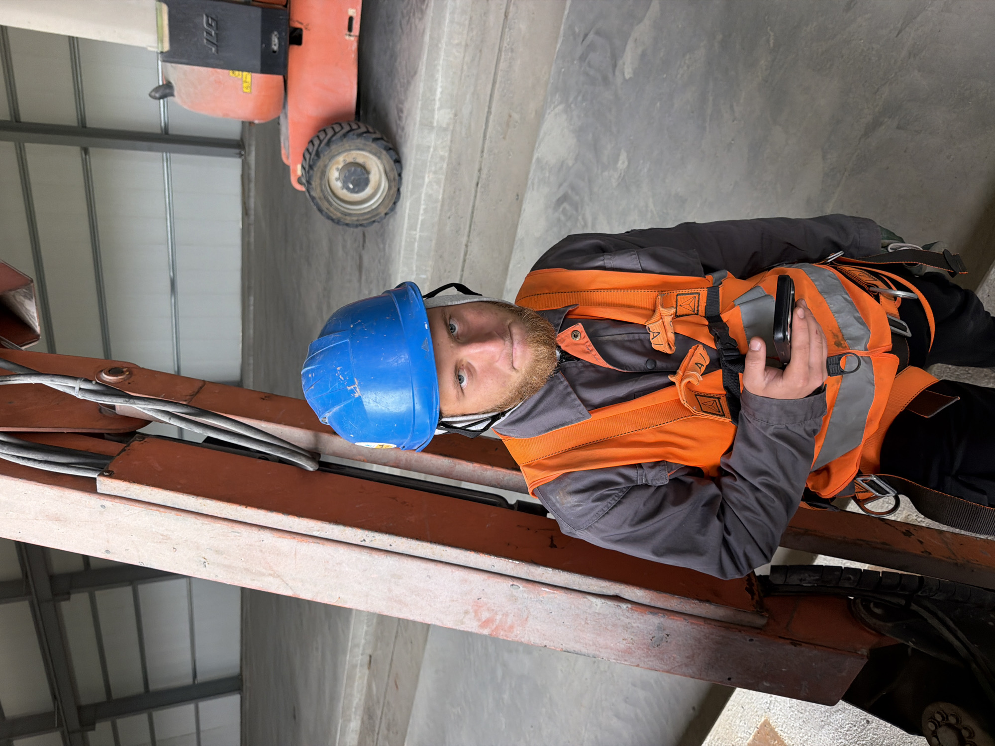An outdoor, medium-shot, eye-level, portrait-oriented photograph captures a young Caucasian male construction worker, approximately in his early 20s, standing in front of an orange hydraulic lift arm. He is wearing a blue hard hat, a dark grey work jacket, and a bright orange safety vest with reflective silver stripes. The vest is equipped with a safety harness, featuring black straps and silver buckles. In his right hand, he holds a black smartphone. The man has light skin, short brown hair, and a reddish-brown beard and mustache. His facial expression appears serious as he looks directly at the camera.
The background reveals an industrial setting, likely a construction site or warehouse in Stróża, Poland, characterized by a concrete floor and the large, grey metal beams and white paneling of a building structure. To the right of the man, a portion of a red and black aerial work platform (lift) is visible, with its tire resting on the concrete. The lighting suggests it is daytime, with natural light illuminating the scene. The overall impression is one of a worker on a job site, momentarily pausing for a photograph.
