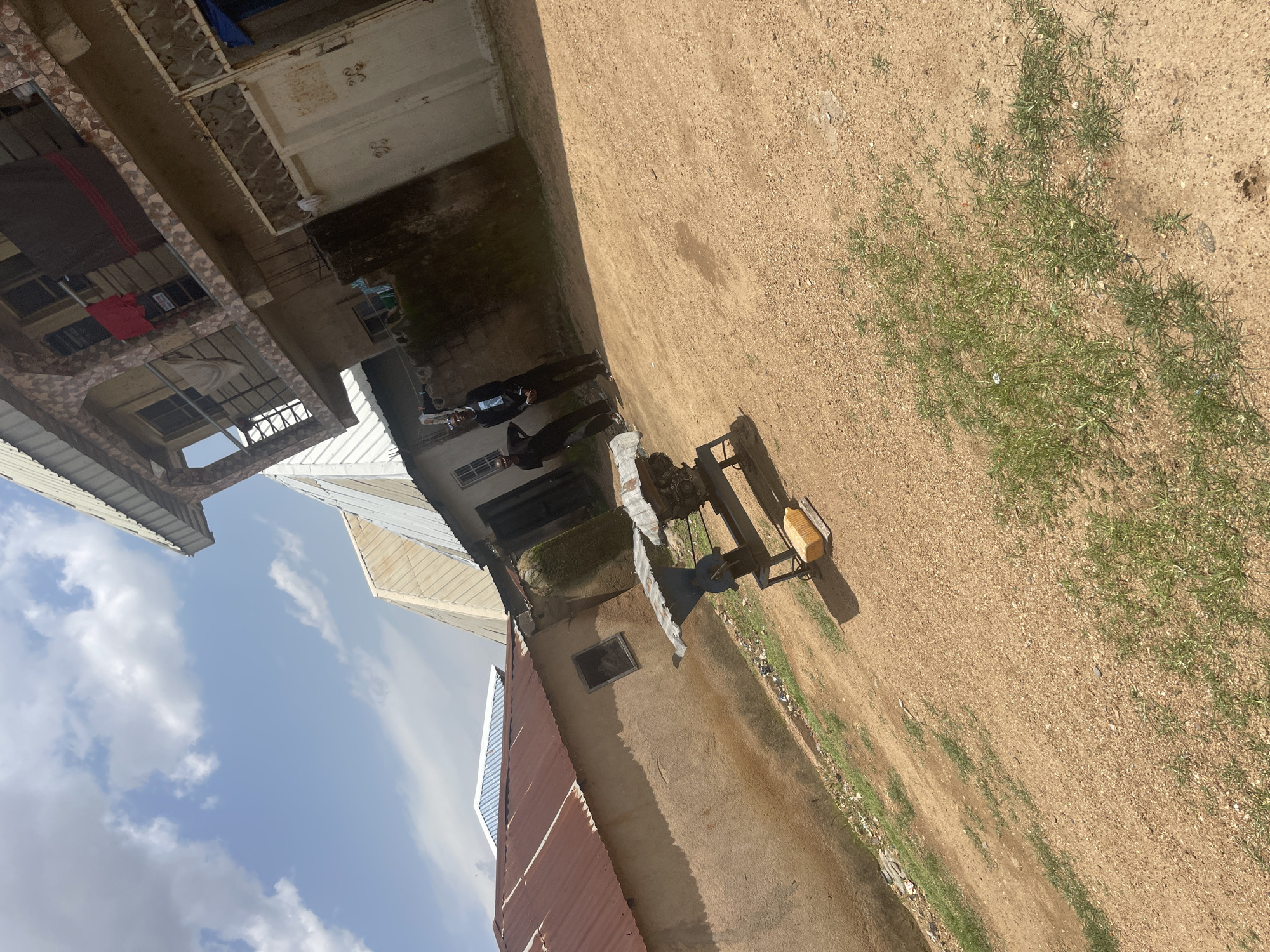 The image depicts an outdoor scene in a residential area in Jos, Nigeria. In the foreground, a dusty, unpaved ground is visible with scattered patches of dry grass. A metallic structure, possibly a piece of machinery or a food processing equipment, stands on a stand. A yellow object, resembling a loaf of bread, is placed on the ground near the machinery.
In the middle ground, two individuals are standing. The person on the right is wearing a dark suit with a jacket and pants, and dark shoes, and appears to be looking towards the camera. The person on the left is wearing a dark t-shirt and dark pants, and is also looking in the general direction of the camera.
Behind the individuals, a building with a textured, multi-colored facade is visible. It appears to be a multi-story structure with balconies and some clothing hanging from a clothesline. To the left of this building, another building with a corrugated metal roof is partially visible. The sky above is partly cloudy with a bright blue sky. The overall environment suggests a typical urban or semi-urban setting in Jos.