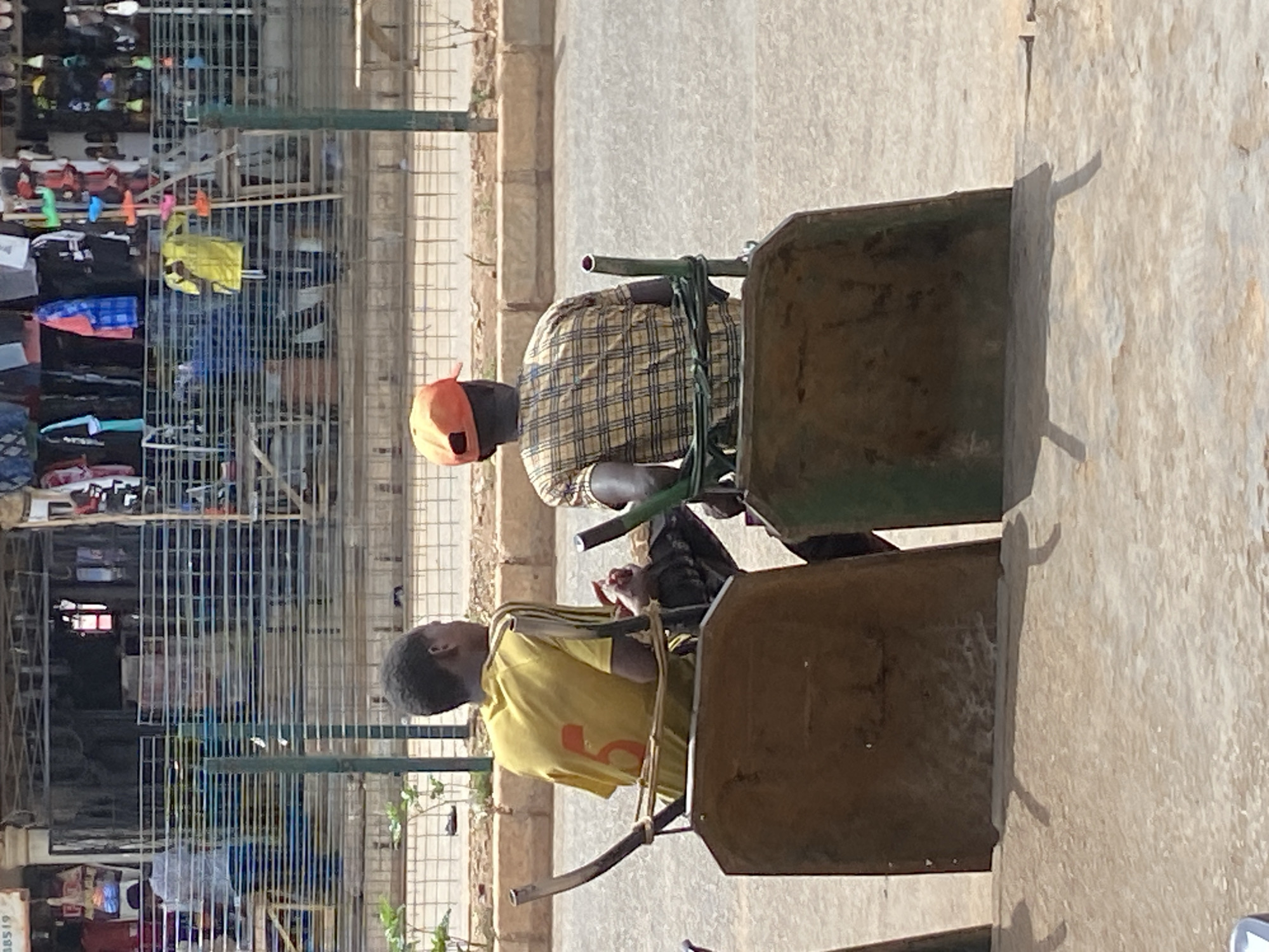 The image, captured in Bukuru, Nigeria, depicts an outdoor scene under bright daylight, evidenced by the strong, distinct shadows cast on the ground. Two individuals, both men, are visible in the foreground, seated on what appear to be repurposed or makeshift seats made from large, sturdy wheelbarrows.
The man closer to the viewer wears a yellow short-sleeved t-shirt with a prominent orange number "5" printed on the back. He has dark, short hair and is looking downwards. His hands are near his lap, possibly holding a small object. He is seated on a dark, metal wheelbarrow with a visible handle frame.
Behind him, another man is seated in a similar fashion, on a dark wheelbarrow. He is wearing a long-sleeved plaid shirt with a pattern of light brown and dark checks, and an orange baseball cap. He is facing slightly away from the camera. Green straps or ropes are visible around the handle area of his wheelbarrow.
The background reveals a bustling market environment. Numerous metal frames and racks are laden with various goods, primarily clothing items in different colors (including yellow, blue, and black). These stalls suggest a street market setting where vendors display their wares. The ground throughout the scene appears to be unpaved, possibly dirt or concrete, consistent with an outdoor market area.
The overall impression is one of people resting or waiting in an active market area during the day. No clear text or signage, other than the number "5" on the shirt, is discernible within the visible scene.