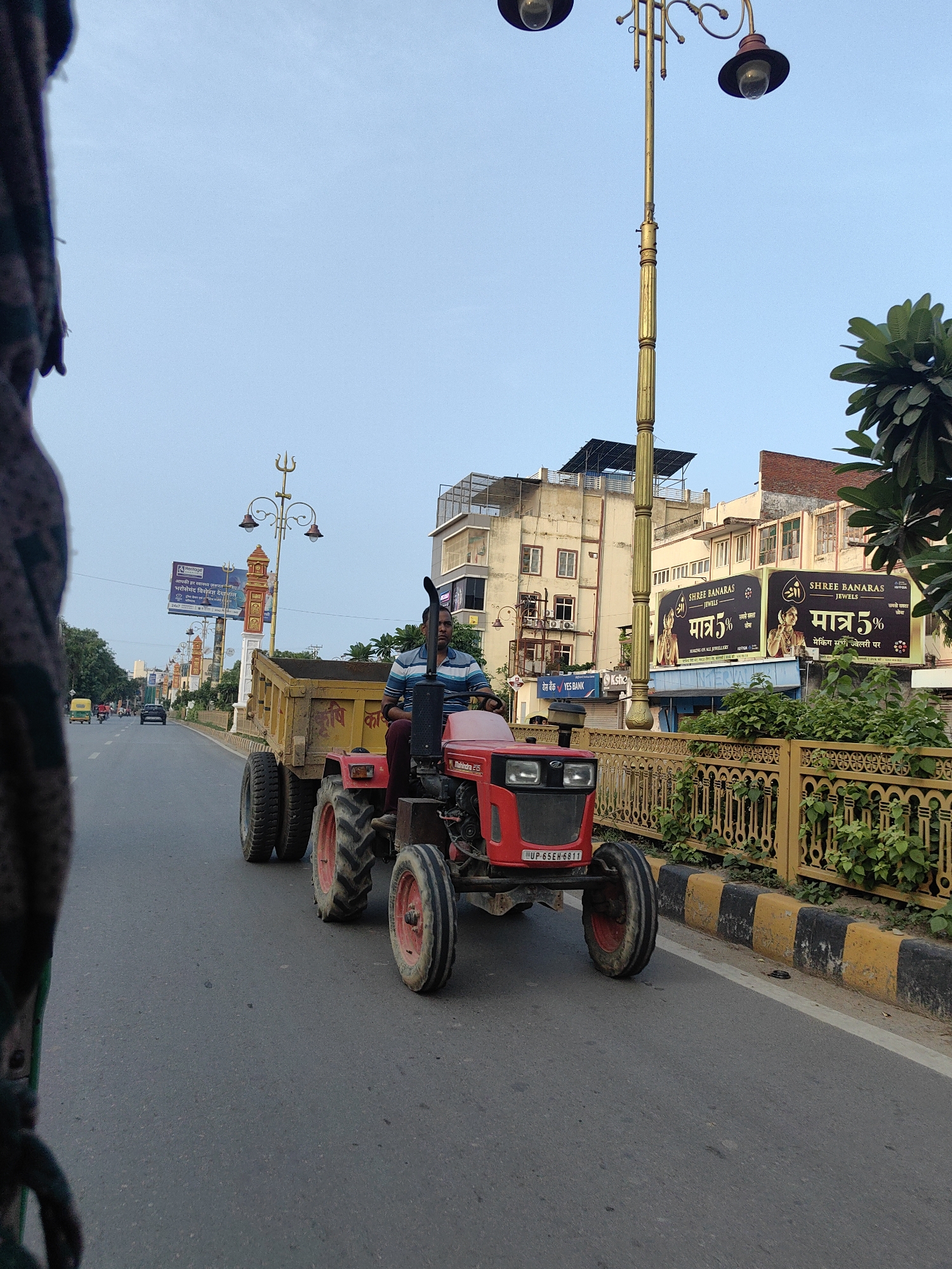 A red Mahindra 215 tractor, driven by a man wearing a blue and white striped shirt and dark pants, is on a paved road in Varanasi, India. The tractor is pulling a yellow trailer. The man appears to be looking forward. The tractor is on the right side of the road, and there is traffic in the distance, including a yellow auto-rickshaw and a black car. Buildings are visible on the left and right sides of the road, with billboards on the right advertising "SHREE BANARAS JEWELS". A decorative yellow fence lines the right side of the road, with greenery behind it. A street lamp with two lights is visible in the background. The sky is a clear blue.