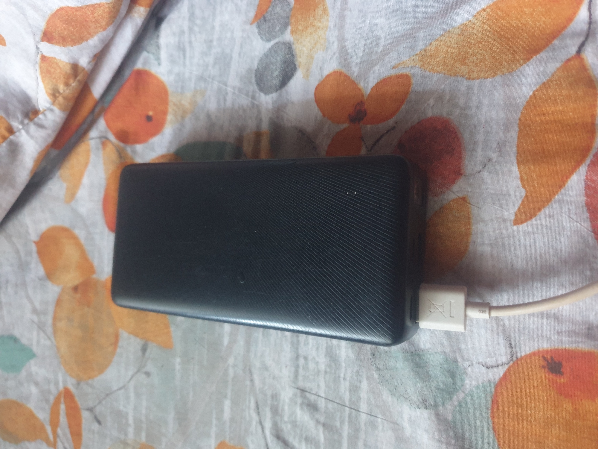 A black, rectangular power bank with rounded corners and a diagonal ribbed texture on its visible surface rests on a light-colored fabric. The power bank shows signs of minor surface wear, including faint scratches and dust. A cream-colored USB-A cable is plugged into the bottom end of the device. The cable's connector housing features an embossed "crossed-out trash can" (WEEE) symbol and small text "020". The fabric background exhibits a repeating pattern of abstract orange, reddish-orange, and faint teal/green leaf-like shapes and irregular grey-black lines on a light grey/off-white base. The fabric appears slightly wrinkled. The scene is static, devoid of human presence or overt action. No discernible identifying features are present to determine a specific location within the city of Unknown, Unknown; the setting appears to be an indoor, likely domestic, environment.