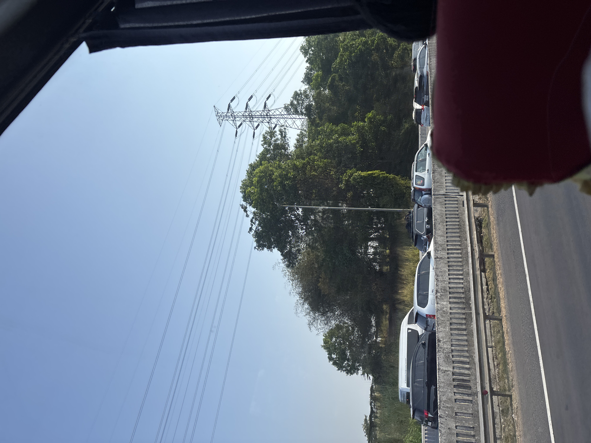 The image captures a traffic jam on an elevated highway in Karawang, Indonesia, likely during daylight hours under a clear blue sky. A series of parallel power lines stretch across the upper portion of the frame, supported by a tall electrical pylon visible in the middle ground. Dense green foliage, including trees and shrubs, borders the highway. On the highway itself, numerous vehicles are stationary or moving slowly, including a white van with "PT Brimaksa Sengkurung Transport" visible on its side, a black SUV, and several silver cars. The perspective suggests the photo was taken from inside a vehicle, with a portion of a red seat in the foreground. The highway appears to have a concrete barrier separating the lanes and a metal guardrail on the outer edge.