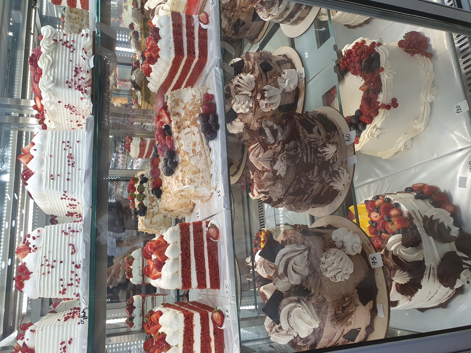 This is a high-angle, eye-level, full shot of a bakery display case filled with various cakes in Batumi, Georgia. The display case is illuminated from above, creating reflections on the glass. The cakes vary in size, shape, and decoration, with many featuring white frosting, berries, and chocolate accents. Some of the cakes are whole, while others are sliced and displayed as individual portions. Price tags are visible on some of the cakes, with numbers like "35", "75", "60", and "120" legible. The background is a blurred view of more bakery items and the interior of the establishment. There are no people clearly visible in the image, though shadows suggest the presence of individuals behind the display. The overall impression is one of abundance and a wide selection of sweet treats.
