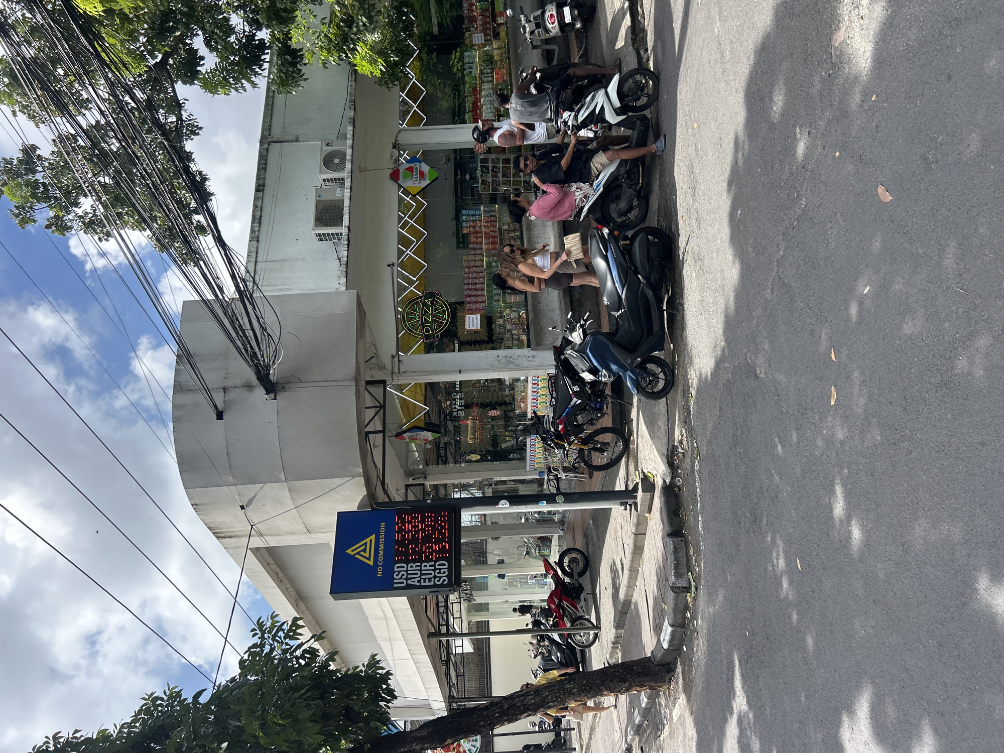 The image depicts a street scene in North Kuta, Indonesia, on a sunny day with scattered clouds. In the foreground, a paved road with dappled sunlight is visible. To the left, a sidewalk borders the road, lined with a tree and a curb. A prominent feature on the sidewalk is a digital currency exchange board displaying exchange rates for USD, EUR, AUD, and SGD, with the text "NO COMMISSION" at the top.
In the middle ground, several motorcycles and scooters are parked along the sidewalk. Some individuals are interacting near the motorcycles. A woman in a yellow shirt and shorts is walking away from the camera on the sidewalk. Further down the sidewalk, a group of people, including men and women, are gathered around motorcycles.
Behind the motorcycles and people, a white building with a curved facade occupies the background. The building appears to be a commercial establishment, with large glass windows revealing shelves stocked with goods. Signs visible on the building include "PIZZA" and "HUNGRY." Several air conditioning units are mounted on the top of the building. A complex network of electrical wires hangs overhead, crossing the sky and partially obscuring the building and trees. The overall environment suggests a commercial area with a lively street presence.