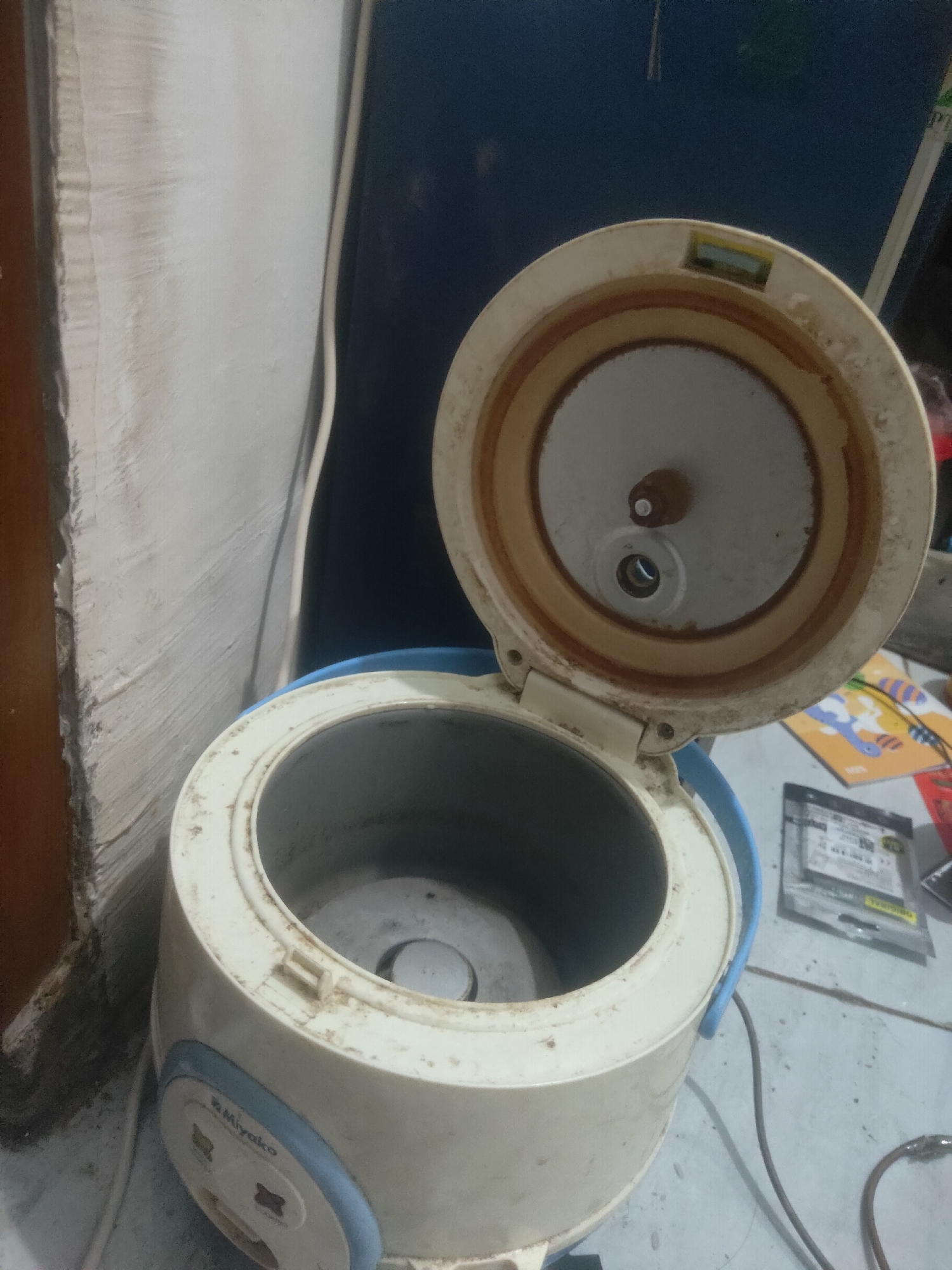 A used, off-white and blue Miyako brand rice cooker is shown from a slightly elevated, three-quarter perspective. The lid of the rice cooker is open, revealing the interior cooking pot and the underside of the lid. The rice cooker is positioned on a light gray floor with a white wall and a dark blue surface in the background. Several electronic items and a small package are visible on the floor to the right of the rice cooker. The scene is indoors, likely in a residential setting in Antirogo, Indonesia.