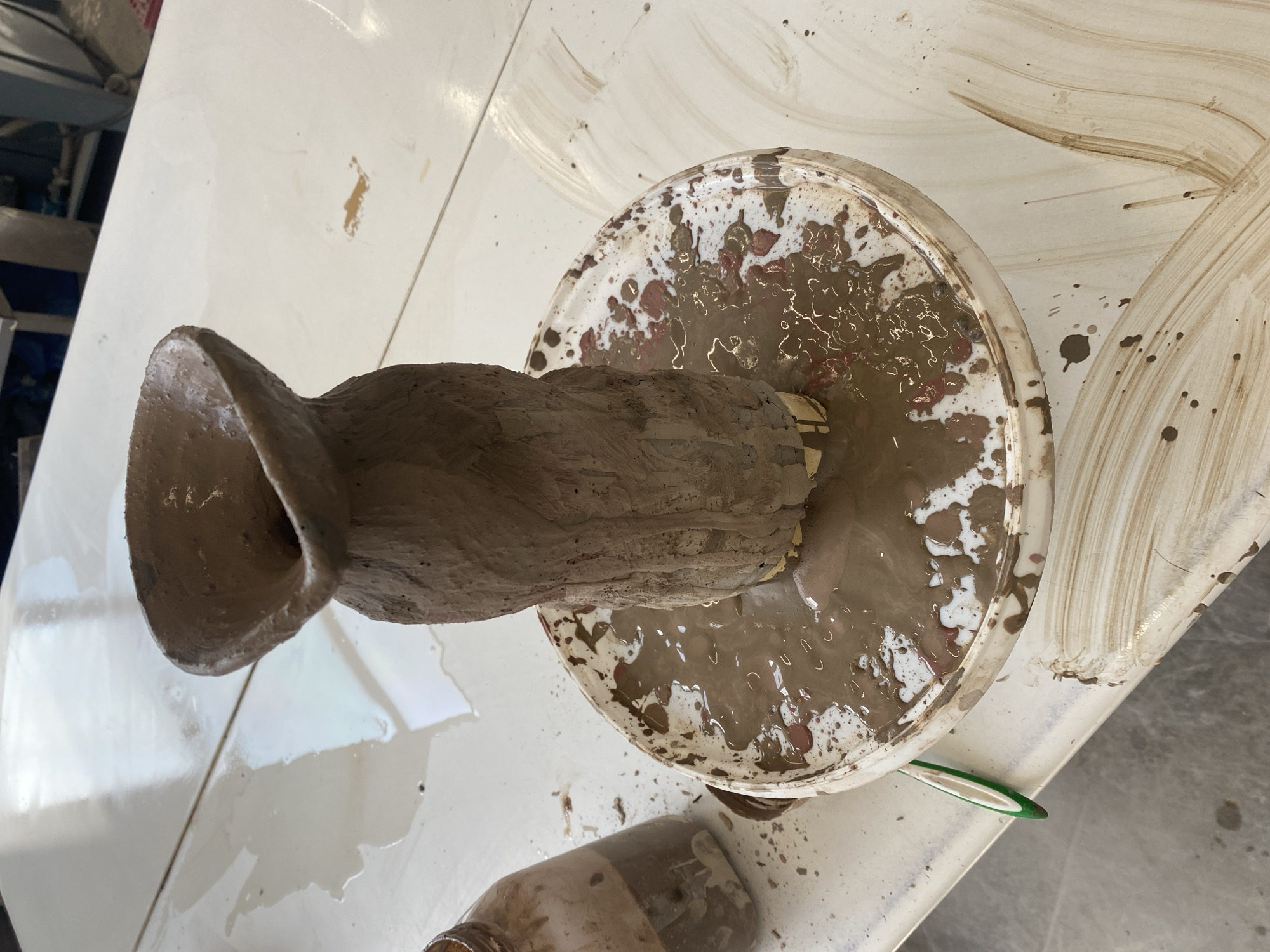 The image shows a clay sculpture on a pottery wheel, likely in a studio setting. The sculpture is rough and appears unfinished, with a wide, open top. The wheel is covered in mud and clay residue. The environment includes a white table covered in splashes and a glass jar. The location is not discernible from the image.