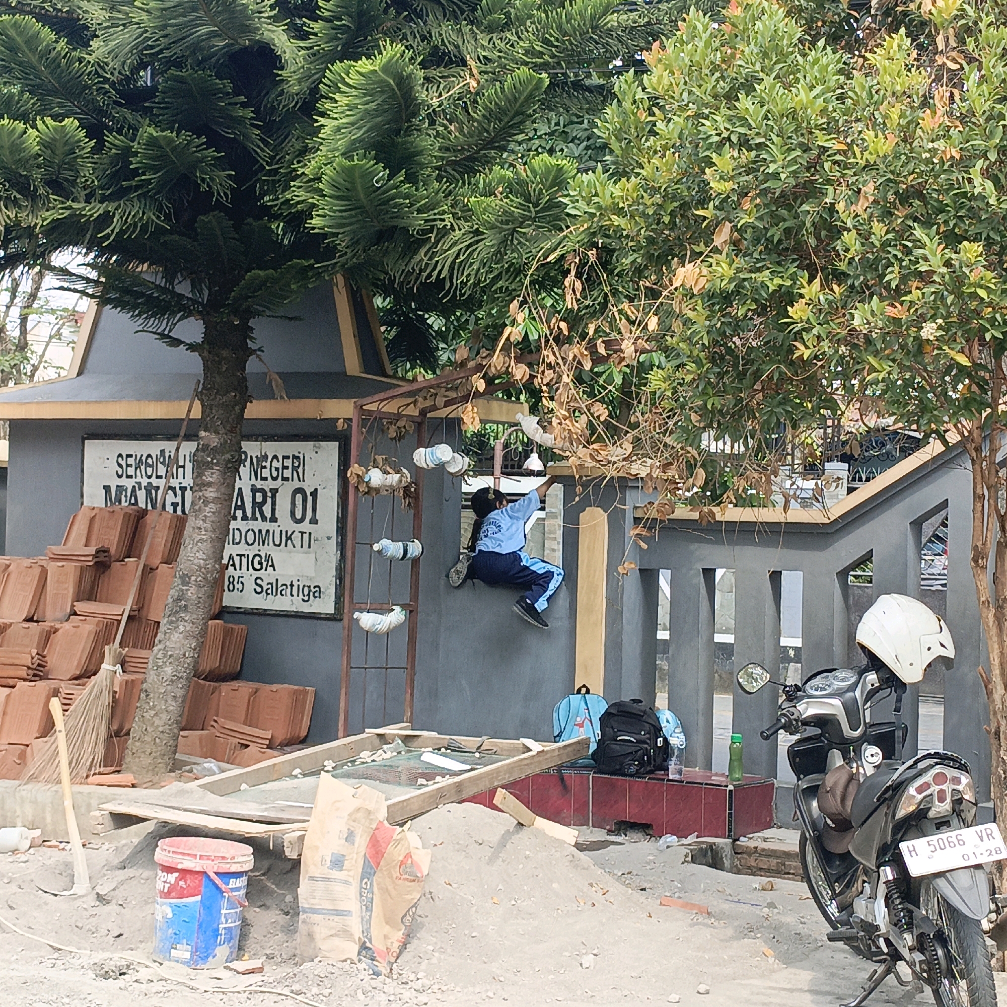 A child is climbing on a decorative metal structure in front of a building with a sign that reads "SEKOLAH NEGERI MANGGUHARJO 01" and lists an address in Salatiga, Indonesia. The building appears to be undergoing renovation, with stacks of terracotta tiles and piles of sand and cement visible in the foreground. A motorcycle with a white helmet is parked near a grey fence. Green trees and foliage surround the scene.