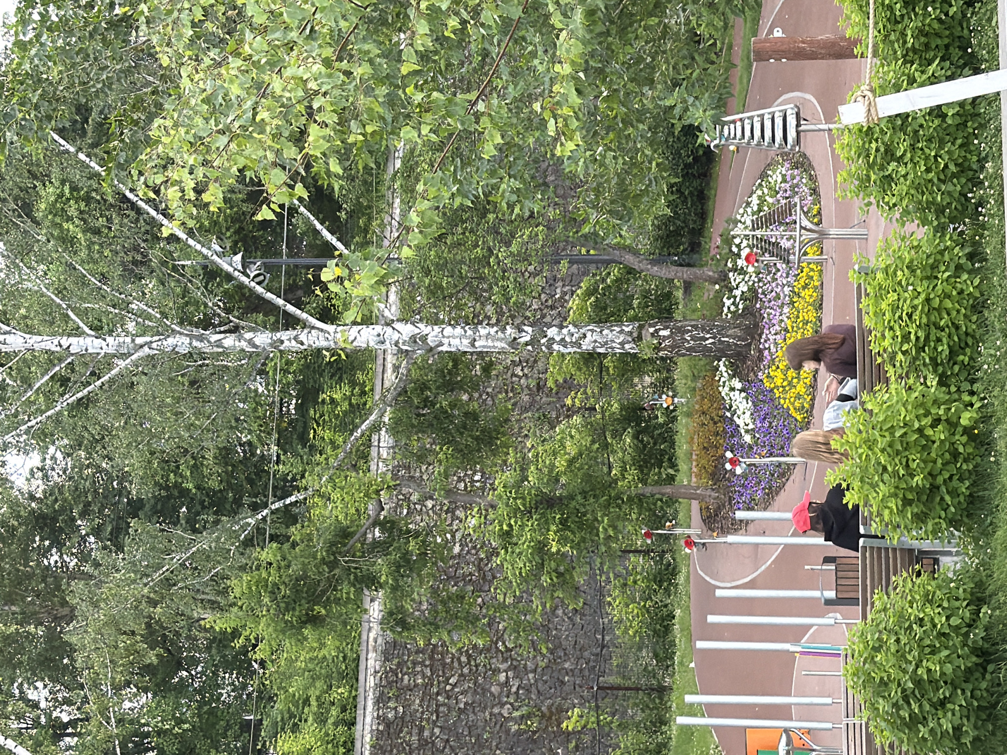 In this outdoor scene, two people are seated on a bench, looking at their phones. The setting is a park with a walking path, a flower bed filled with colorful blooms, and lush green trees. One person is wearing a dark top and a red baseball cap, while the other is wearing a dark top and has long blonde hair.
A tall birch tree with white bark stands prominently in the foreground. In the background, a stone wall is visible, suggesting a possibly historical or architecturally interesting park. The ground is a reddish-brown color, likely a synthetic surface for a recreational area. Several metal pipes, resembling a musical installation or a part of a playground, are also present. Small decorative elements, like red and white ladybug figures, are scattered around the flower bed.
The image appears to be taken during the daytime, with overcast skies suggested by the diffused lighting. The overall atmosphere seems peaceful and relaxed, with the individuals engaged in quiet activities amidst nature. The location context of Perm, Russia, doesn't provide further specific visual cues in this image. No visible text is present.