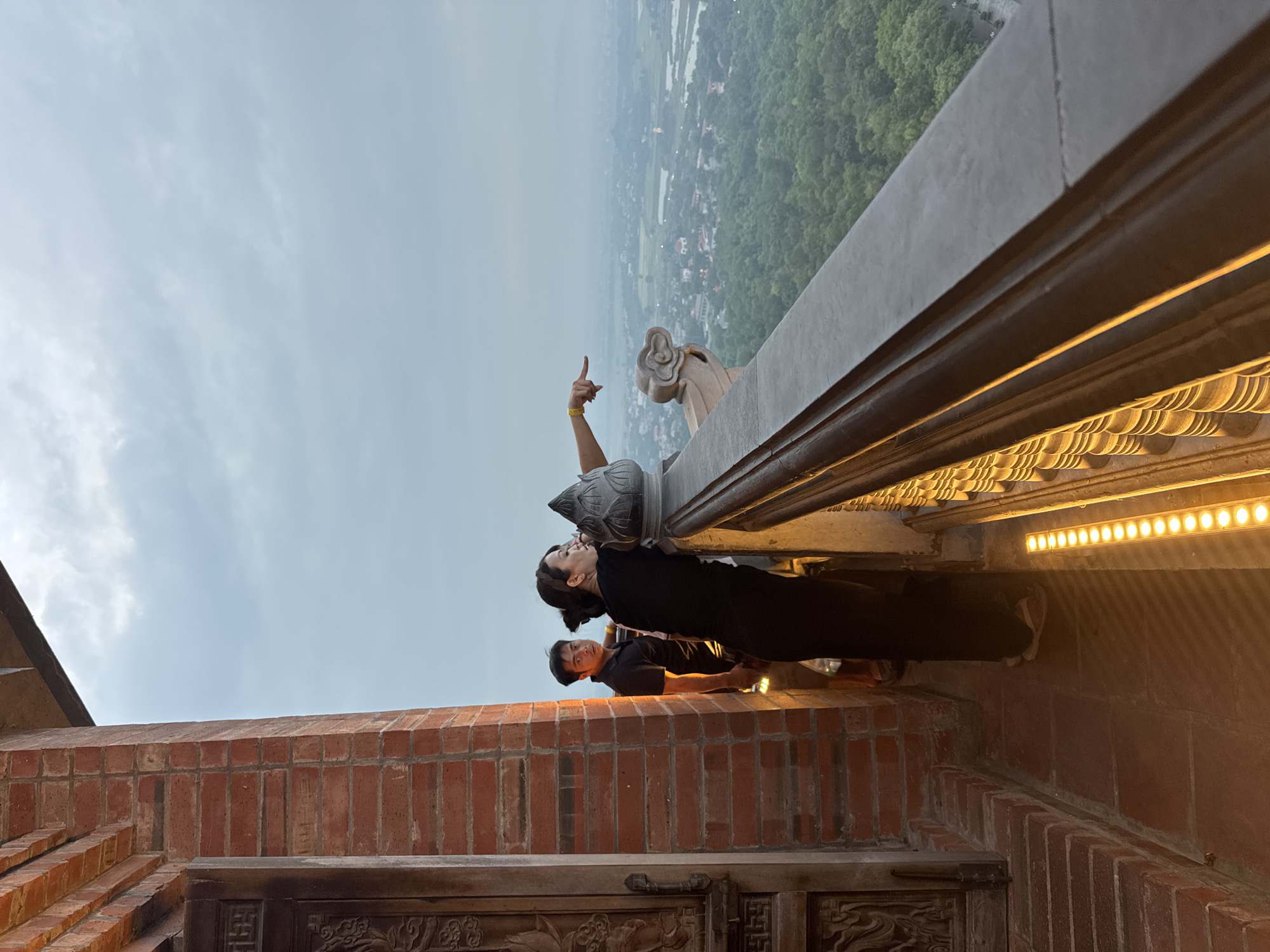 This image captures a view from a vantage point in Tây Hoa Lư Ward, Vietnam, likely from a temple or a high structure. In the foreground, a brick pillar and a carved wooden door are visible on the left. A woman, dressed in black, is leaning on a railing and pointing towards the expansive landscape. Beside her, a man in a black shirt looks towards the camera. The railing is decorated with ornate carvings and illuminated by a strip of yellow lights. Below the railing, a lush green forest stretches out, interspersed with fields and water bodies, with a hazy city in the distance. The sky above is overcast with clouds.