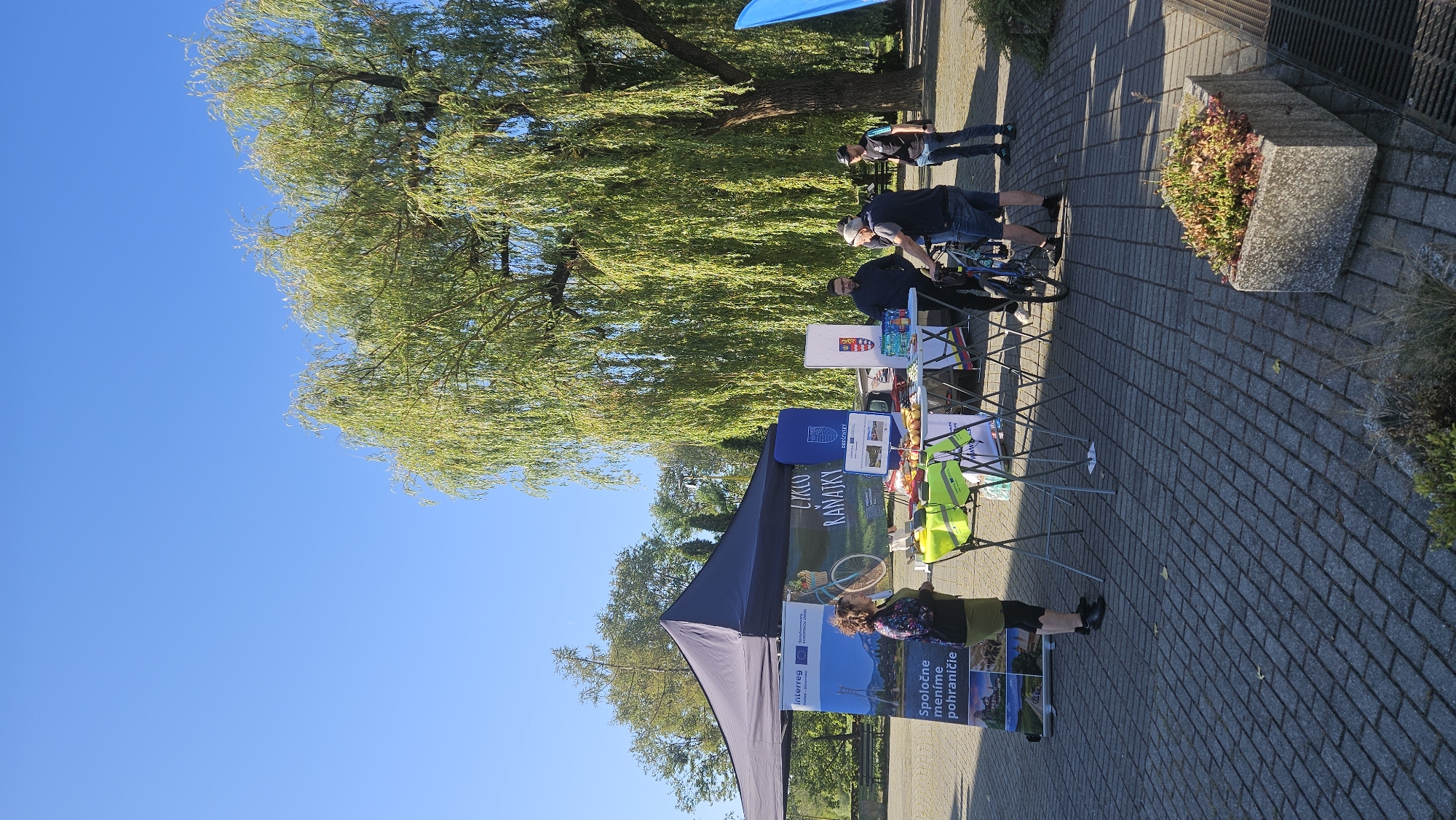 The image depicts an outdoor event under a large, mature willow tree, possibly in the city of Prešov, Slovakia. The scene is dominated by a dark blue tent, behind which several people are gathered around tables displaying promotional materials. A banner on the left side of the tent features the Interreg and European Union logos, along with text in Slovak which translates to "Together we are changing the border". Another banner with text "LYKKO RANAJKY" and a bicycle illustration is visible.
Several individuals are interacting at the tables. One woman, wearing a green skirt and a patterned top, stands near the banner. Three men are visible near the tables, one of whom is leaning over a table, while another stands with his arms crossed. A bicycle is parked near the tables.
The ground is paved with grey interlocking bricks. In the foreground, a concrete planter filled with plants is visible. The sky is clear and blue, indicating a sunny day. The overall atmosphere suggests a community event or information fair, potentially related to cycling tourism or cross-border cooperation, given the visible branding and imagery.