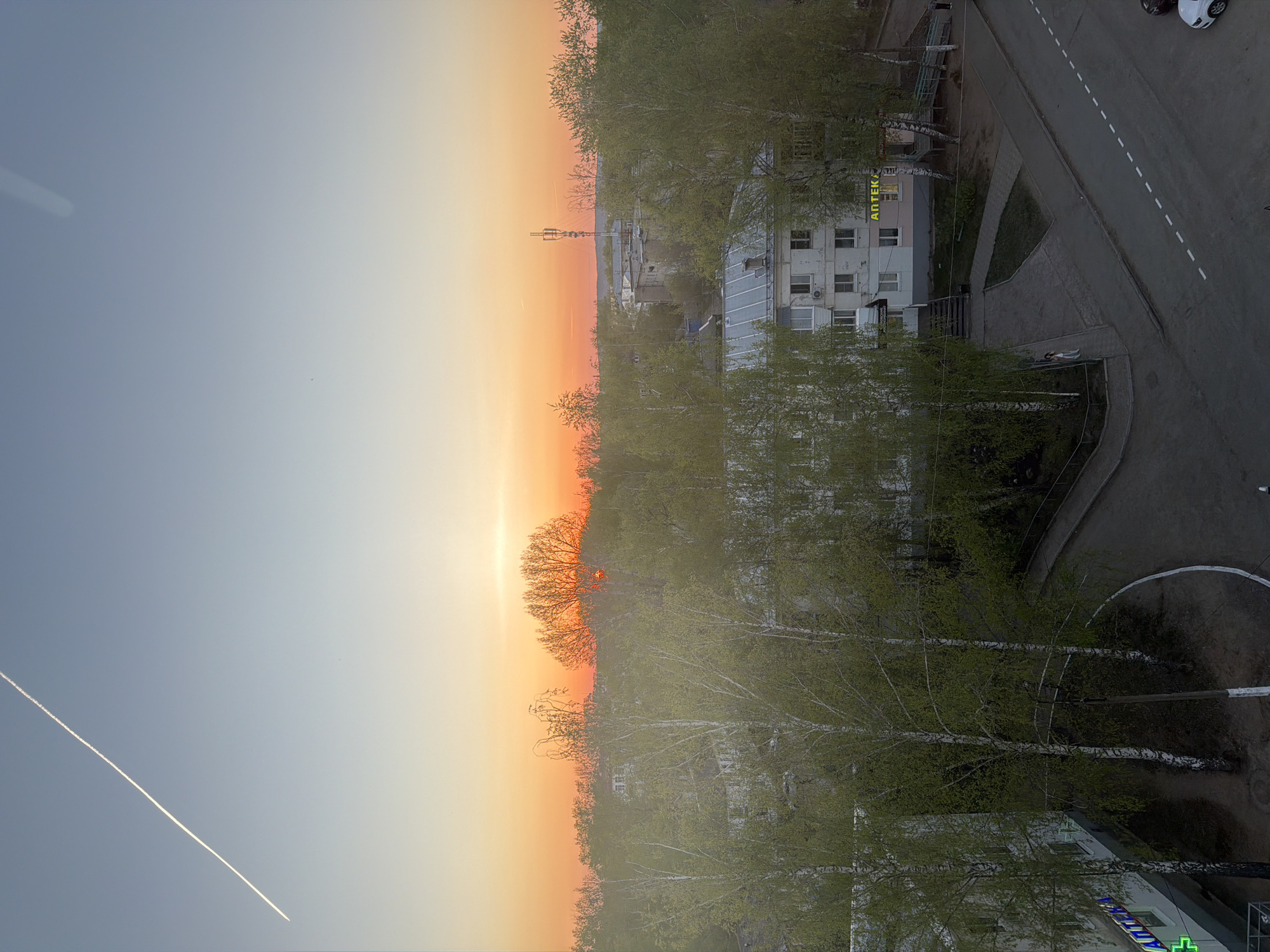The image shows a sunset view from a high vantage point in Chaikovsky, Russia. The sky transitions from blue to orange as the sun sets. A contrail from an airplane is visible in the sky. Below, the skyline is lined with trees, with some bare branches and others with new green leaves. A building complex is visible, including one with a sign in Russian that reads "АПТЕКА", indicating a pharmacy. A road runs adjacent to the building, with white dashed lines marking lanes. A couple of parked cars are visible on the road. The overall scene depicts an urban environment at sunset.