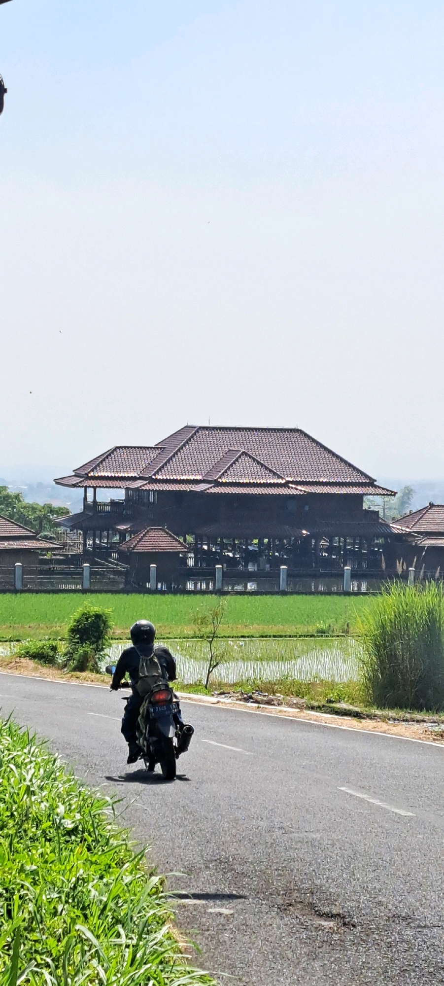 A medium shot captures a motorcyclist from behind, riding on a paved road that curves to the right, surrounded by lush green vegetation on the left and a bright, hazy sky above. The motorcyclist, wearing a black helmet and backpack, is positioned in the lower left quadrant of the frame, their motorcycle facing away from the viewer.
In the middle ground, a large, dark wooden structure with a prominent, multi-tiered tiled roof stands prominently. This building appears to be an open-air venue, possibly a restaurant or guesthouse, with visible wooden posts supporting its upper levels and balconies. Adjacent to this structure, a vibrant green rice paddy stretches out, reflecting the sky. The entire scene is set against a backdrop of distant, hazy hills.
The location is identified as Watuagung, Indonesia, suggesting a rural or semi-rural setting with traditional architecture and agricultural landscapes. The atmosphere is bright and sunny, indicative of daytime.