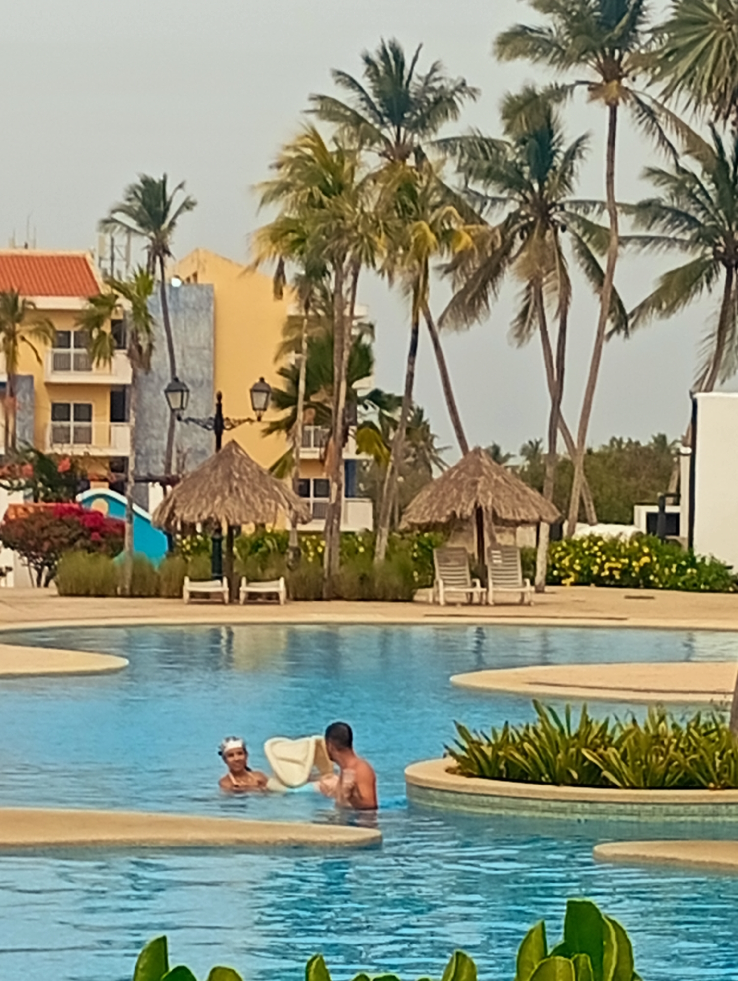 This image was taken in Playa El Agua, Venezuela. It shows a swimming pool in a tropical resort setting. Two people are in the pool, one wearing a white swim cap. A man stands near the person with the swim cap holding a white pool float. In the background, there are palm trees, thatched-roof huts, lounge chairs, and a multistory hotel building with balconies. There are green plants and flowers around the pool and resort area. The sky is overcast.