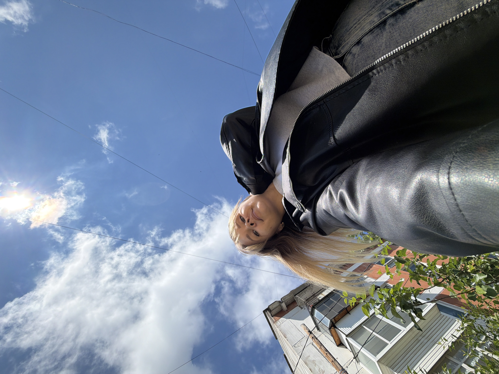 This is an extreme low-angle, wide-shot selfie taken outdoors in Chaikovsky, Russia, on a bright, sunny day. The main subject is a young woman with long, straight blonde hair, visible from her upper torso upwards. She is looking downwards towards the camera with a neutral to slightly smiling expression. She wears a black leather jacket over a light grey top, partially obscured by the jacket's lapel and collar.
The camera's perspective is pointed upwards, making the vast blue sky with scattered white cumulus clouds a dominant feature of the image. A strong sun flare is visible in the top-left corner, indicating clear weather. Several thin power lines or wires traverse the sky horizontally and diagonally.
In the bottom right of the frame, the upper part of an old, light-colored building is visible, featuring windows and what appears to be a balcony or architectural detail. Green foliage from a tree or bush extends into the right side of the frame, partially obscuring the building. The building's facade shows signs of wear and age. The overall scene captures an outdoor urban or suburban setting from an unusual, dynamic perspective. There is no visible text in the image.