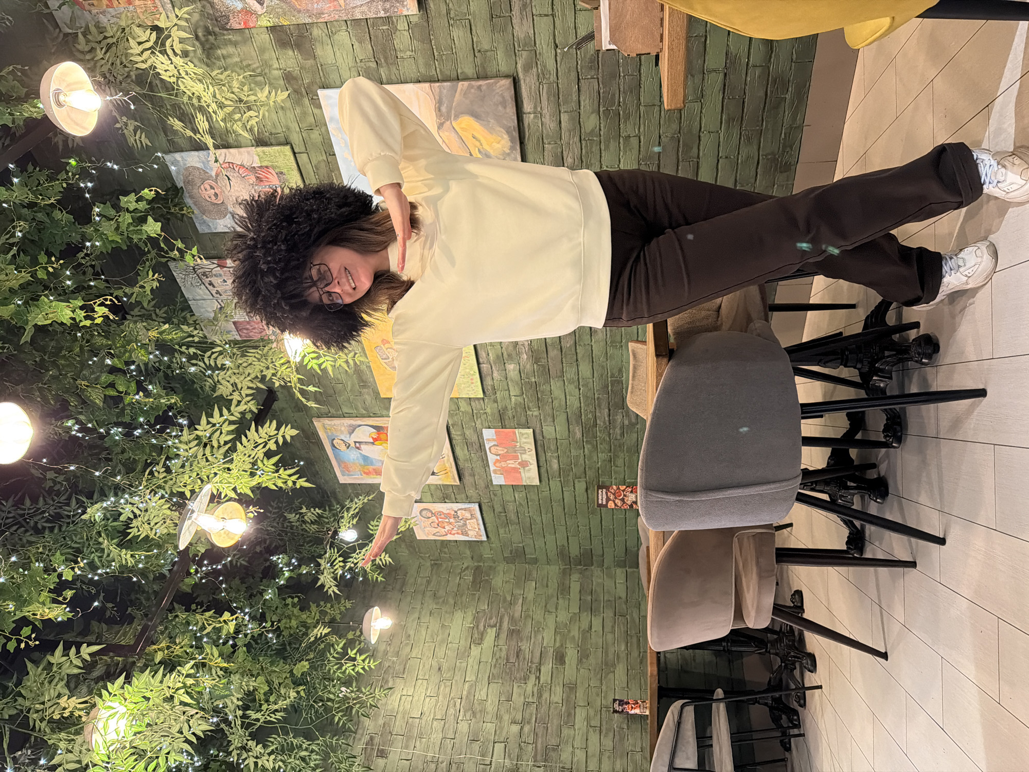 The image shows a woman wearing glasses and a large, dark, fluffy hat, standing in a space decorated with artificial foliage and string lights. She's wearing a light-colored sweatshirt and dark pants, with white sneakers on her feet. She's gesturing with both hands, one up and one down. The space appears to be indoors, with a brick-textured wall adorned with small paintings or illustrations. There are chairs nearby, and the floor has a light, tiled surface. The location in the city is unknown.