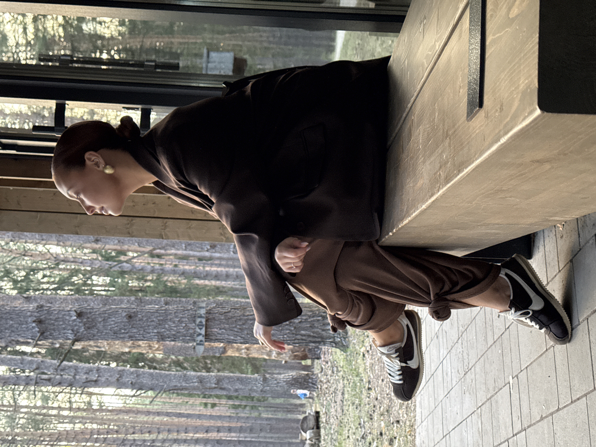 A woman in a brown coat and brown pants sits on a wooden ledge. She has a bun hairstyle and is wearing earrings. She is wearing black and white sneakers. Behind her is a forest view with trees and sunlight. The location is in a city called Votkinsk, Russia, based on a sign on one of the trees that says "ВУФП". The ground is paved with grey tiles.