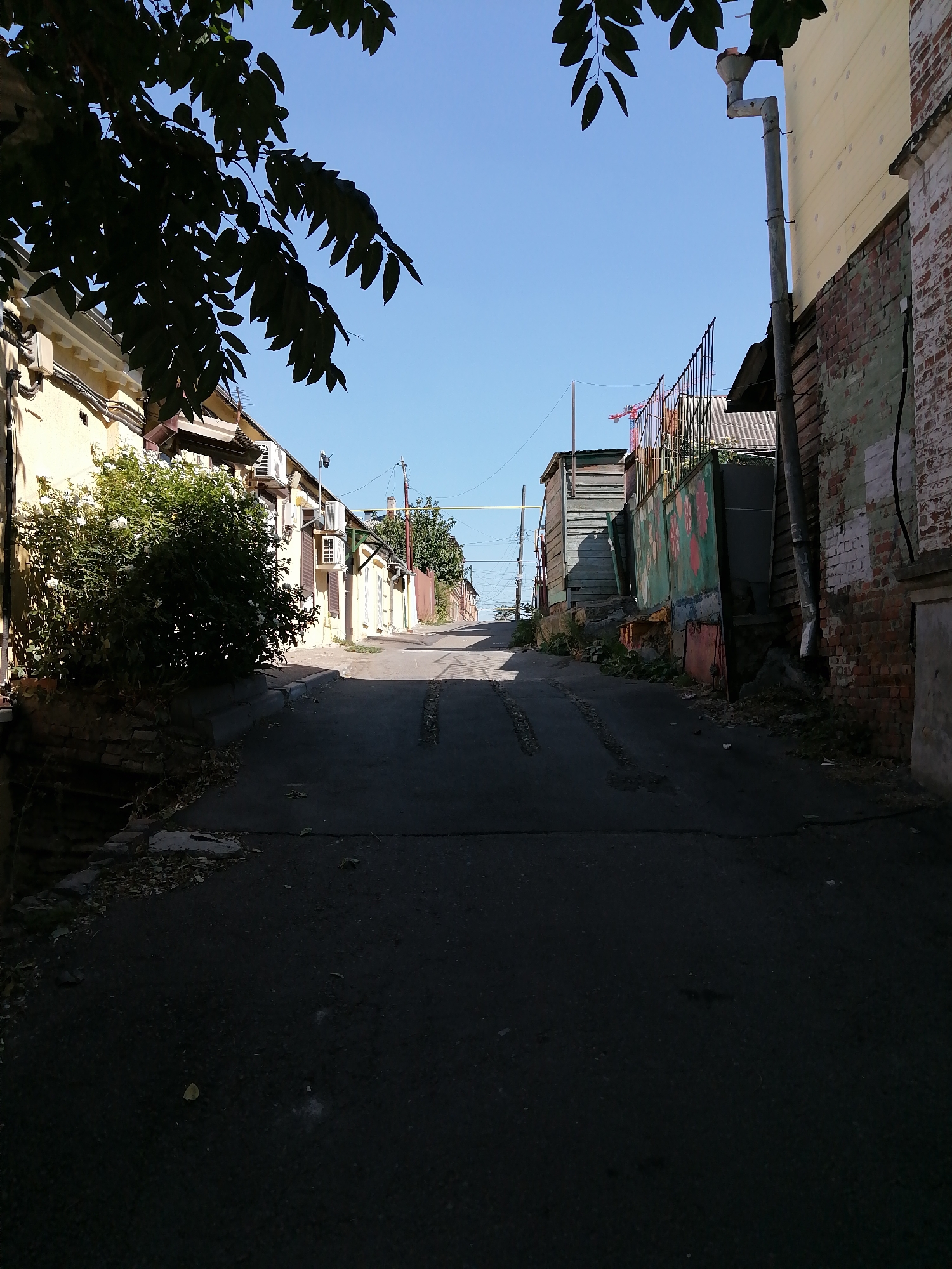 The image depicts a narrow, paved alleyway in Rostov-on-Don, Russia, on a clear, sunny day. The alley slopes upwards and is flanked by buildings on both sides. On the left, a yellow building with a white cornice and visible air conditioning units is partially obscured by a lush green bush with white flowers. A dark, leafy tree hangs over the scene from the top left. On the right, the alley is bordered by a weathered brick wall, and further down, wooden structures and a metal fence with colorful painted floral designs. A construction crane is visible in the distance beyond the fence. The asphalt road shows signs of wear, with some visible tire tracks and scattered debris. The overall impression is of a typical, somewhat aged urban environment.