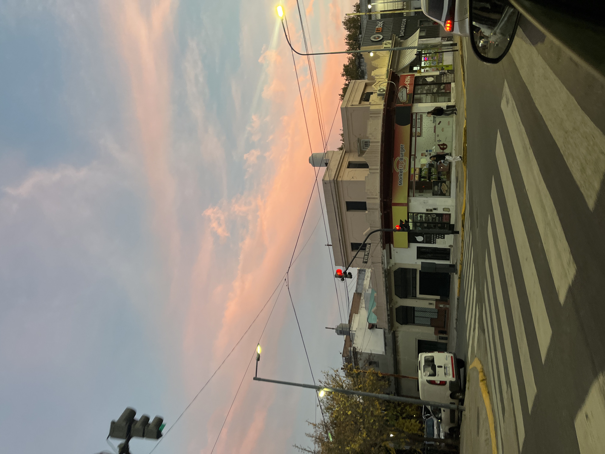 An eye-level view from the street looking at the opposite side of a street. A crosswalk is visible, and several pedestrians are standing in front of a store. Buildings of varying height and design face the street. The sky is pink and blue. Traffic lights show red and green signals. Street lights illuminate the scene. A van and other vehicles are parked or driving on the street. A car mirror is visible in the top right of the image. The location is in the city of Buenos Aires, Argentina, on Eva Perón.