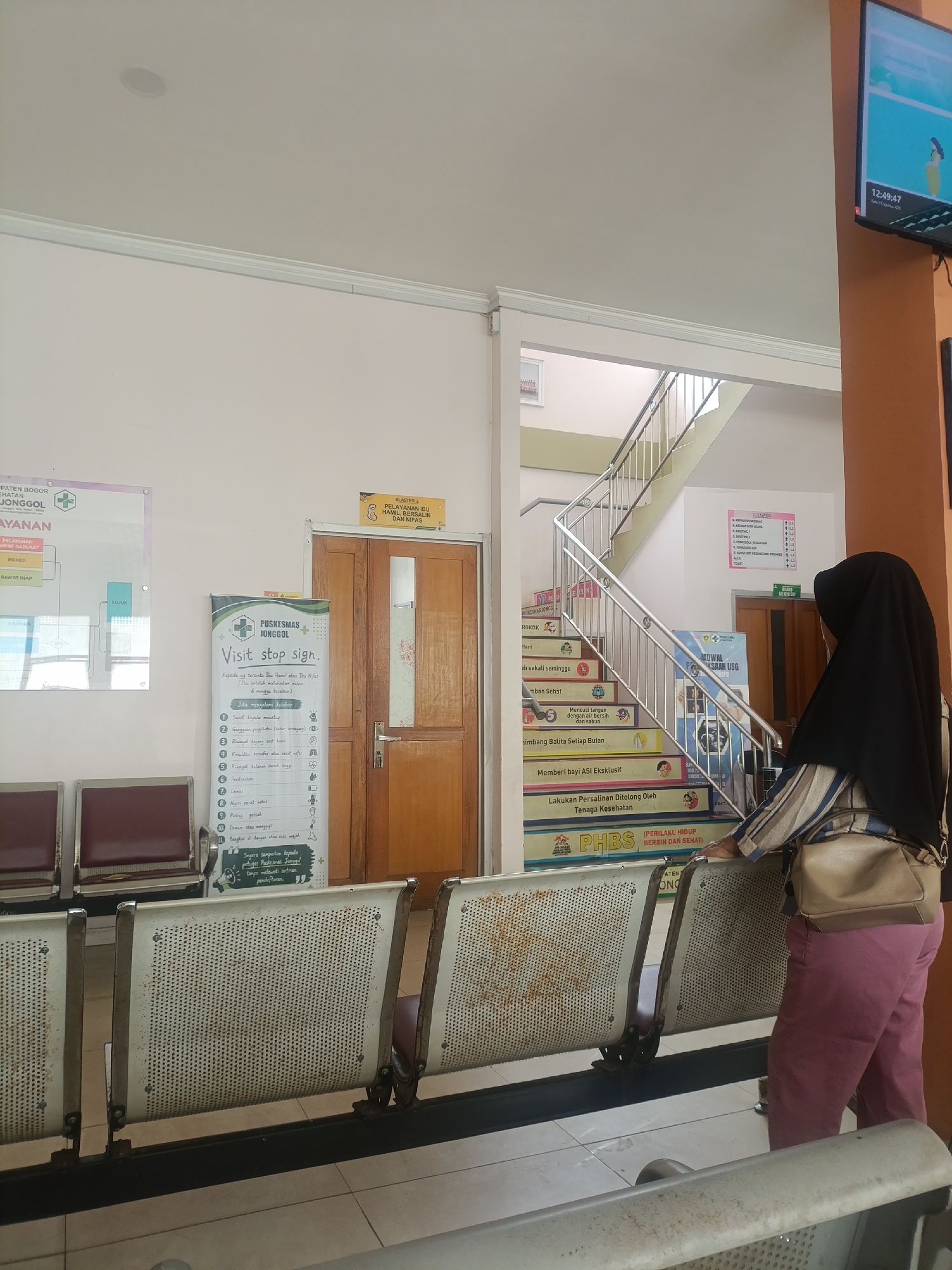 The image depicts the waiting area of a public health center (Puskesmas) in Jonggol, Indonesia. In the foreground, a row of worn, metal bench seats with maroon upholstery are visible. To the right, a woman wearing a black hijab and a striped shirt with beige pants stands and leans on one of the benches. She has a light brown shoulder bag. Behind her, a wooden door with a glass panel is situated next to a staircase. The staircase features colorful posters and informational signs on its steps, promoting health awareness. A prominent poster titled "Visit Stop Sign" is displayed near the door. On the left side of the image, another poster with a green and white logo is mounted on the wall, displaying service information for the health center. A large television screen is mounted on an orange pillar to the right, displaying a clock showing 12:49:47 and a graphic. The environment suggests a public service facility, likely a clinic or hospital waiting room.
