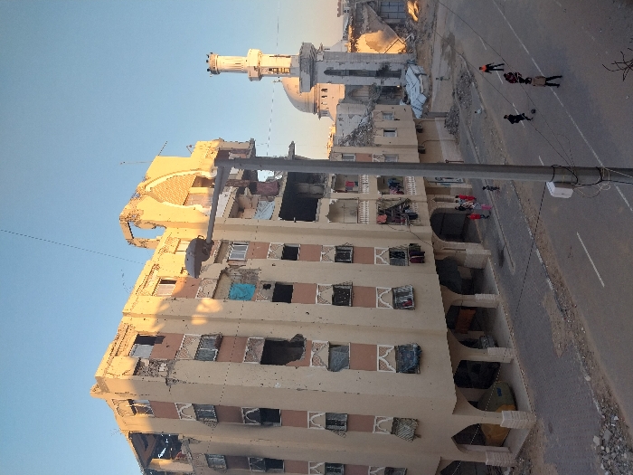 An elevated view captures a street scene in Khan Yunis, Palestinian Territory, characterized by significant structural damage under a clear, warm-toned sky suggesting early morning or late afternoon.
In the foreground, a prominent multi-story residential building, possibly five or six stories, exhibits extensive damage. Its tan-colored facade is pockmarked with numerous holes, shattered windows, and missing sections of wall, particularly on its upper floors and roof where a large portion of the structure has collapsed, exposing interior rooms and debris. Decorative arches support the ground floor, beneath which a yellow vehicle is partially visible, and several individuals, including children, are gathered on a paved area. Laundry hangs from balconies on the building's intact sections, and a blue tarp is visible in one window opening.
To the right and in the mid-ground, a white mosque with a distinctive dome and a tall, cylindrical minaret is visible, also showing signs of structural damage. Other surrounding buildings appear partially collapsed or reduced to rubble.
A paved road with white lane markings runs across the lower half of the image. On the road, approximately five children are actively playing soccer with a dark ball. A utility pole with wires stands prominently in the mid-ground, partially obscuring the view of the street. Debris and rubble are scattered along the roadside and near the damaged structures.