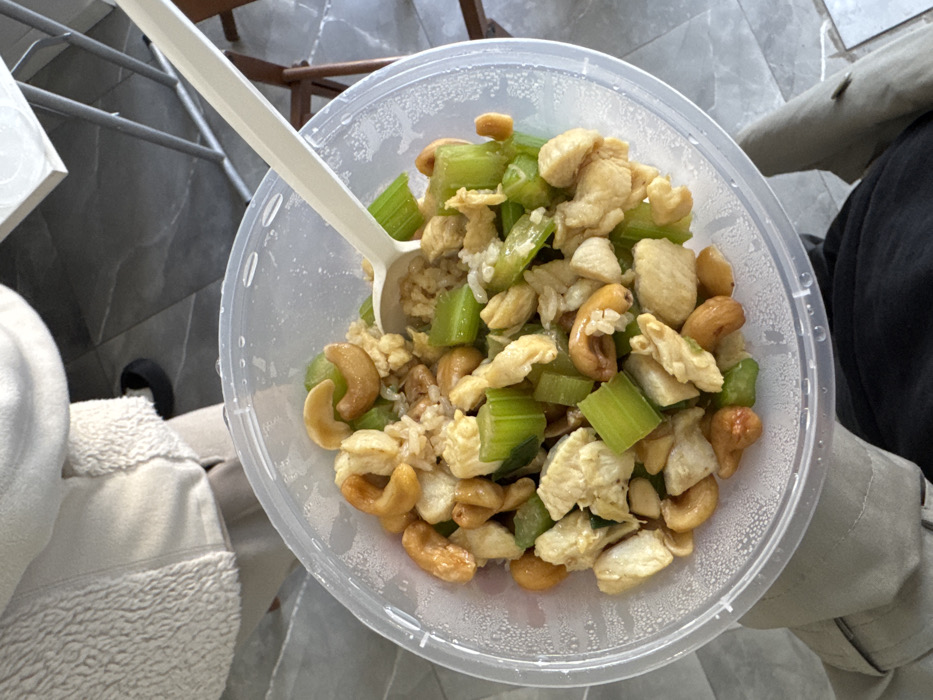 The image displays a clear plastic round take-out container, centrally positioned and held from below by an individual. The container holds a mixed dish consisting of diced cooked chicken, chopped green celery, whole cashew nuts, and grains of rice. A white plastic spoon is inserted into the food. The person holding the container wears dark trousers on the left leg and light beige or grey trousers on the right leg. The upper part of the individual's body, dressed in a cream-colored textured fleece or sherpa jacket, is visible in the upper left. The background features a light grey tiled floor with a geometric pattern. To the right, partial elements of dark brown wooden furniture, including legs and crossbars, are visible. In the upper right, a metallic frame supporting a light-colored surface is partially discernible. The specific location within the city of Milan, Italy, cannot be determined from the visual evidence provided.