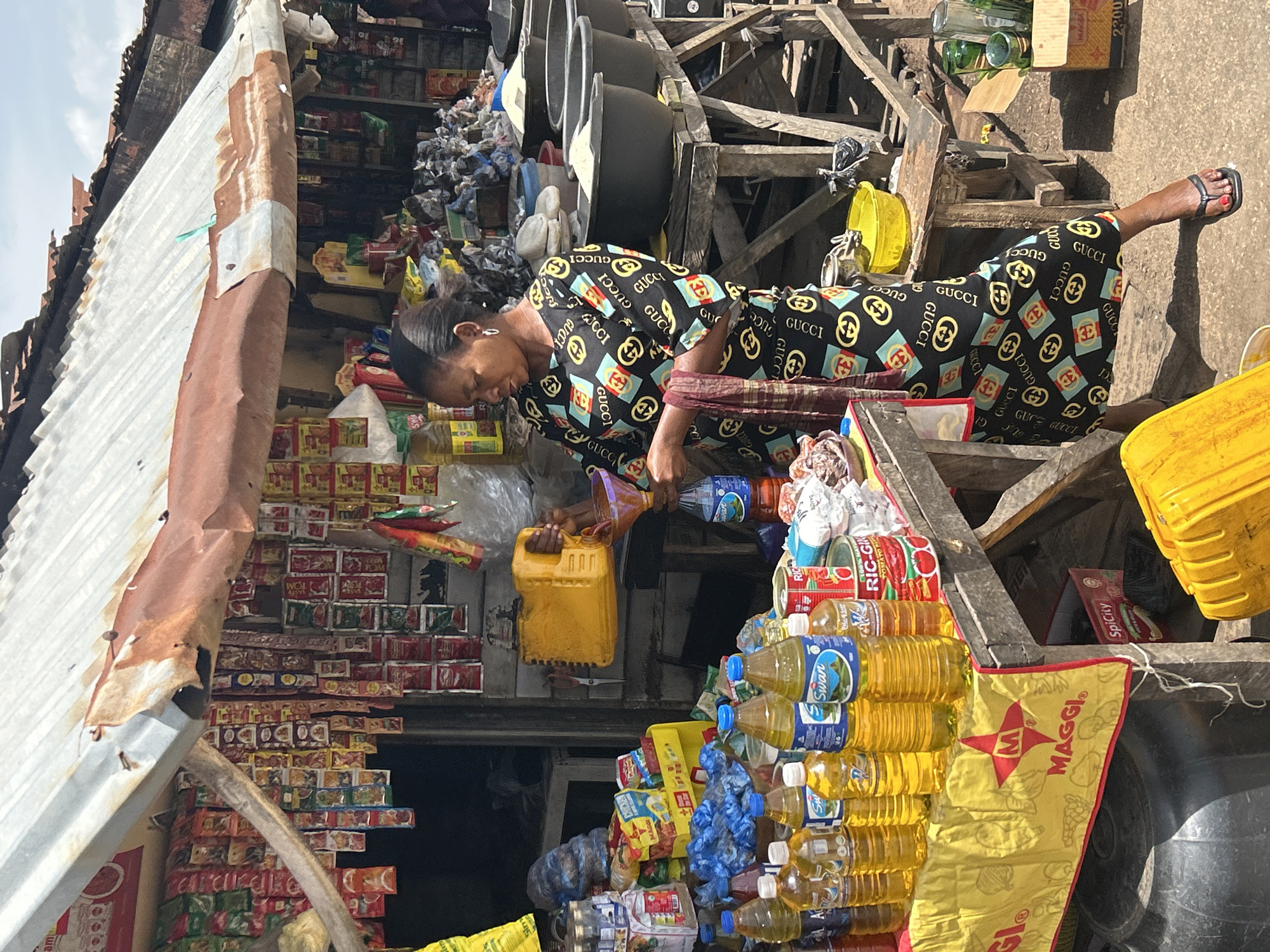 This media file captures a vibrant scene at a bustling market in Jos, Nigeria. The main subject is a woman, dressed in a distinctive black dress adorned with a repeating pattern of interlocking "G" logos and squares in red, blue, and gold, suggestive of a designer print. She is bent over, engaged in an activity that appears to be the pouring of a yellow liquid from a yellow jerrycan into a smaller container. Her head is bowed, and her focused expression suggests an engrossing task.
The setting is a roadside stall, characterized by an abundance of goods. Shelves are packed with various packaged items, including what appear to be food products like seasoning packets and canned goods. Piles of plastic bottles filled with yellow liquid, likely cooking oil, are stacked prominently. A large yellow jerrycan and several other containers, some transparent and others opaque, are also visible. The stall is constructed with a corrugated metal roof and wooden supports, giving it a rustic and temporary feel. In the background, more stacked items and wooden structures suggest the dense and organized chaos typical of a Nigerian marketplace.
The overall impression is one of commercial activity and daily life. The bright sunlight indicates it is daytime, likely mid-morning or afternoon, given the strong shadows. The scene conveys a sense of industriousness and the vibrant energy of a local market. The woman's attire, while eye-catching, is juxtaposed with the utilitarian nature of the market stall and its merchandise, highlighting a blend of personal style and economic endeavor. Text visible on some of the packaging includes "MAGGI" on a yellow banner, and other unidentifiable labels on product boxes and bottles.