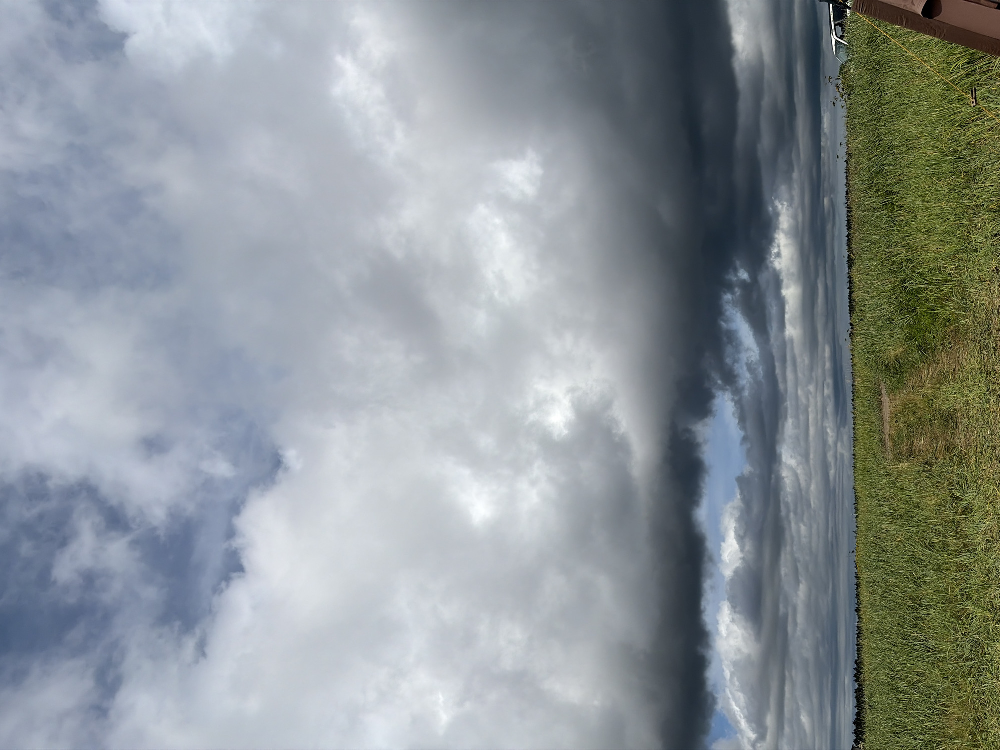 A vast, green field stretches towards a dark, stormy horizon under a dramatic sky filled with heavy, grey clouds. A narrow, dirt path winds through the tall grass, leading the eye into the distance. In the far right of the image, a portion of a dark brown structure is visible, possibly a fence post or part of a building. On the far right edge of the horizon, a white vehicle can be seen amongst the trees. The overall scene suggests a rural setting, possibly on the outskirts of Ozerskoe, Russia, with the approaching storm creating a sense of impending weather.