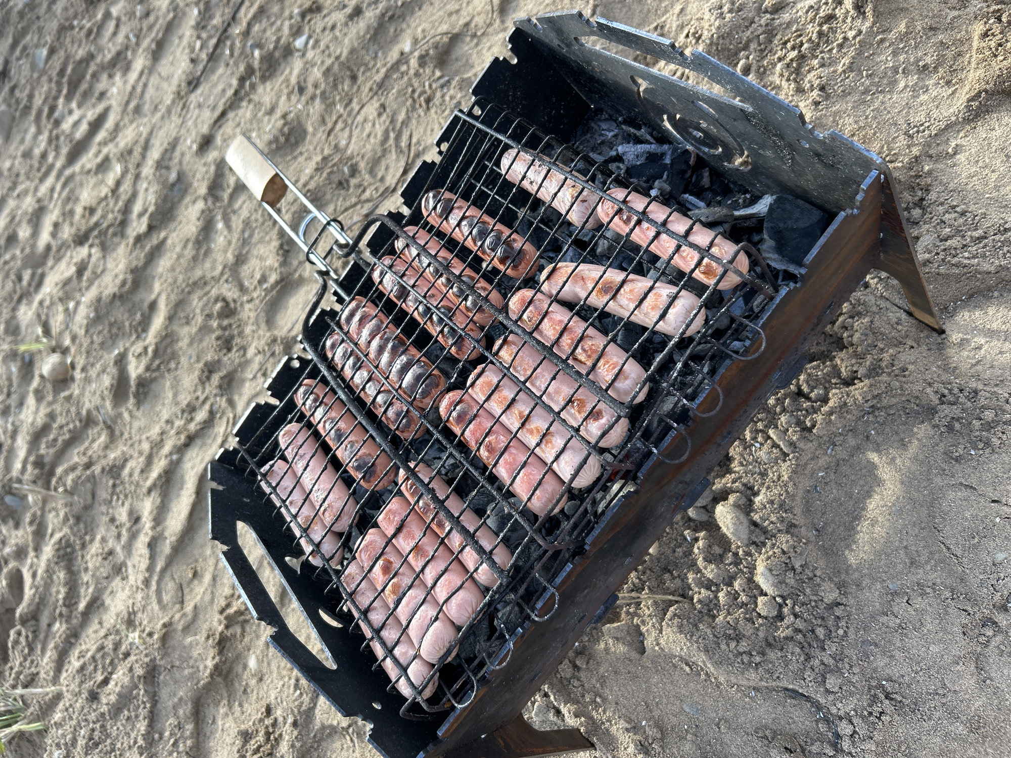 A portable metal grill containing numerous sausages is positioned on sandy ground. Some of the sausages are browning from the heat of the coals beneath. The grill is black and appears to be made of folded metal, with a wooden handle attached to its lifting mechanism. The background consists of light brown sand, showing subtle ripples and scattered small stones and dried grass. No people are visible in the image. The location is the sandy embankment of the Kama River in Chaikovsky, Russia.