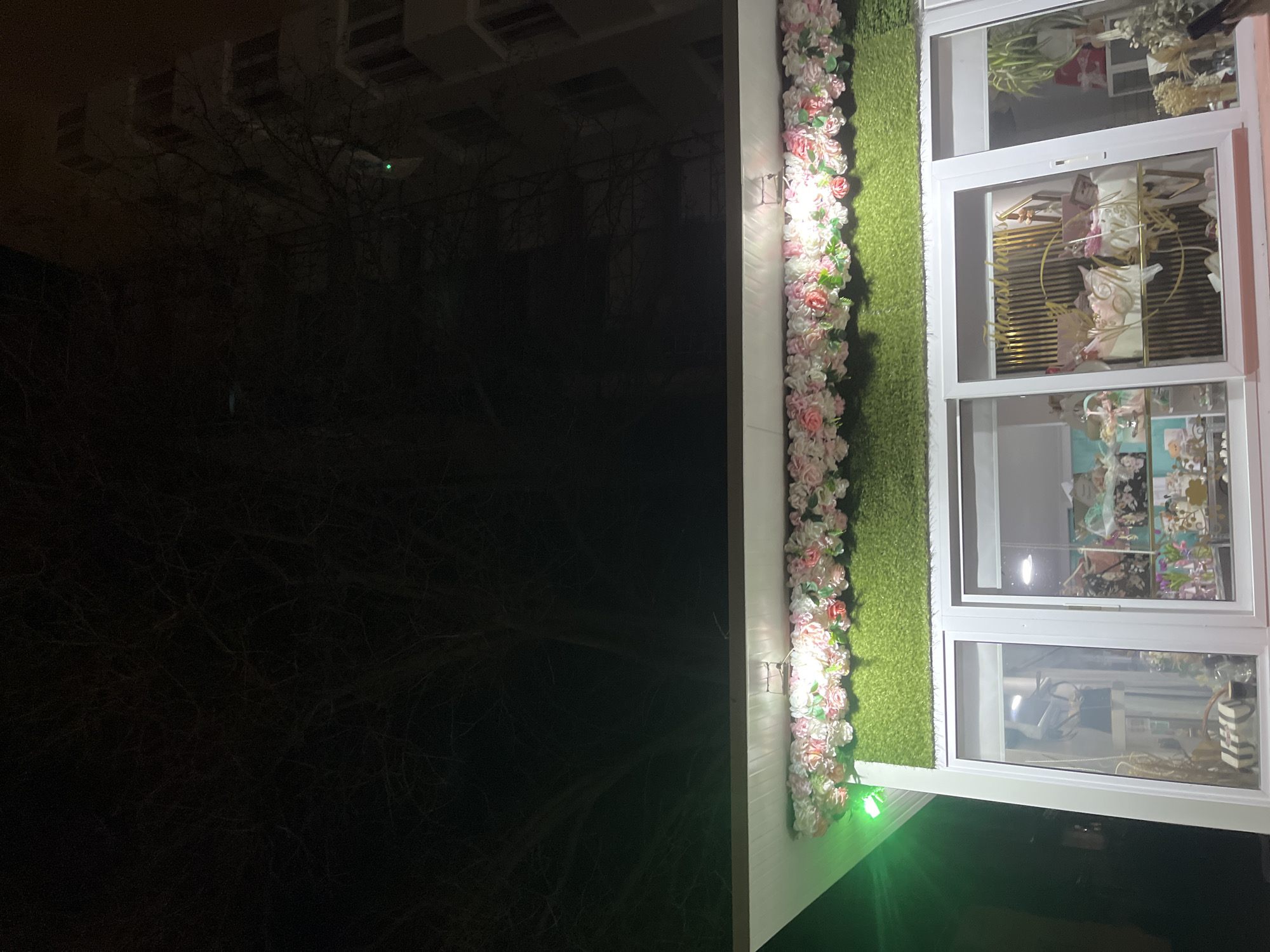 The image shows a shopfront in Goce Delcev, Bulgaria, at night. The shop's exterior is decorated with artificial flowers and a strip of artificial grass. Inside the shop, visible through the window, are various floral arrangements and decorative items. The street is dark, and bare tree branches are visible.