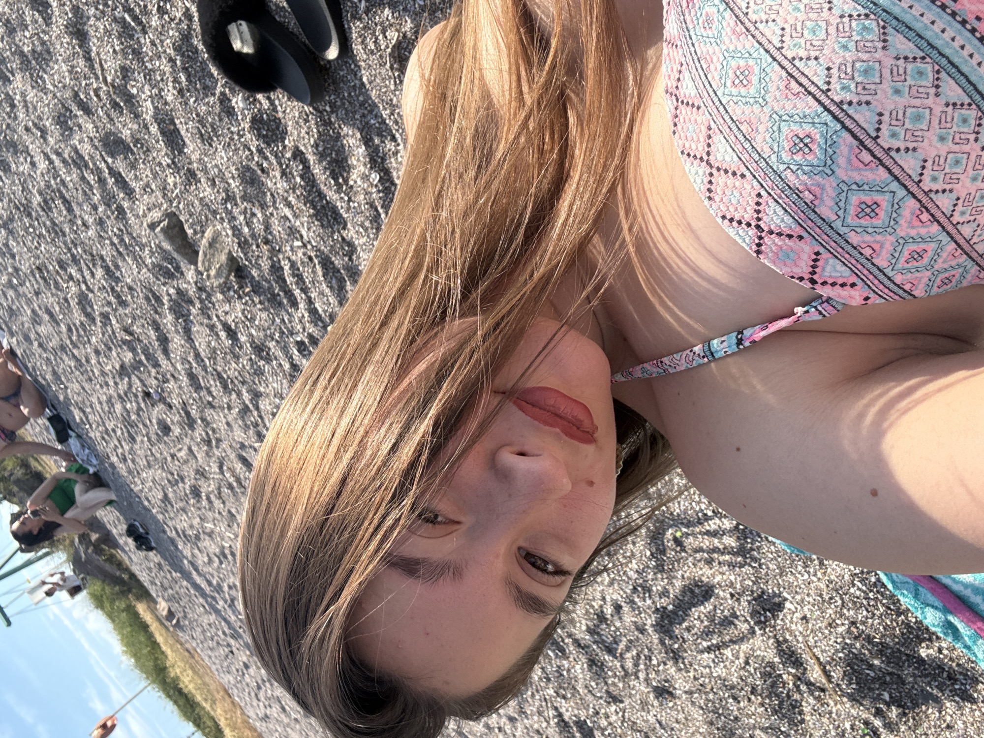 A young woman with long, blonde hair and a patterned bikini top lies on a pebble beach. Her face is turned towards the camera, and she is looking directly into it with a slight smile. Her hair is blowing in the wind. In the background, other people are visible sitting on the beach and one person is on a swing set. The sky is clear and blue. The location is the beach in Sozopol, Bulgaria.