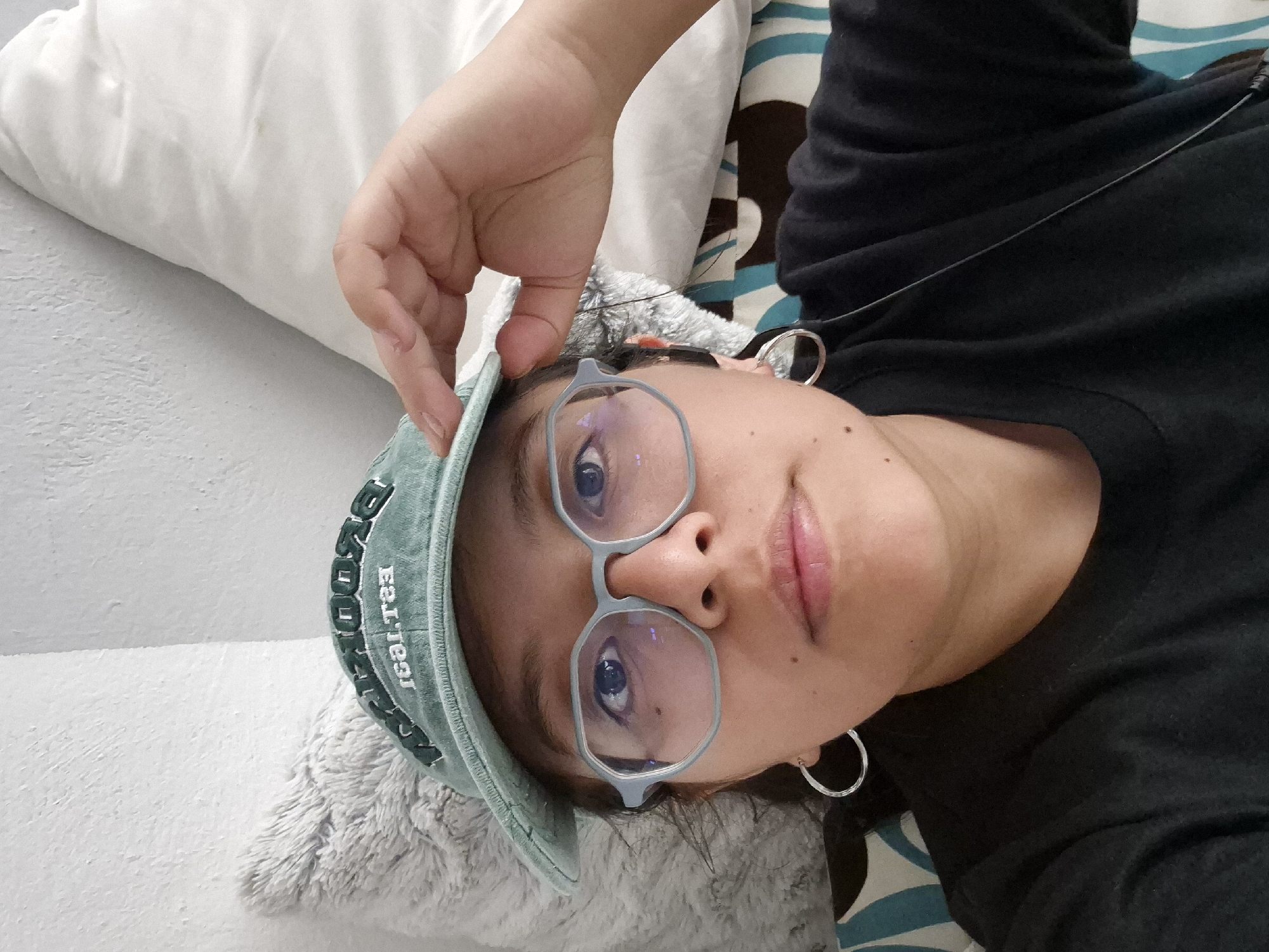 A person is lying down, wearing glasses and a baseball cap. The background includes pillows and bedding with a patterned design. The person is wearing a dark-colored shirt and hoop earrings. Their hand is holding the brim of the cap. The location cannot be determined from the image.