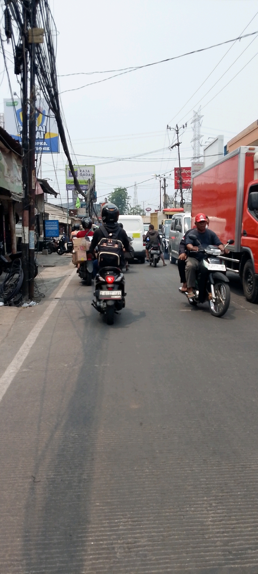 The image depicts a street scene in Bekasi, Indonesia, with a focus on traffic and urban infrastructure. In the foreground, a black scooter with a license plate reading "Z 6137 TAS" is visible, its brake light illuminated. The scooter is positioned on an asphalt road marked with white lines, suggesting a moderately busy street. Behind this scooter, another scooter rider wearing a black helmet and backpack is seen from the rear, also on the road. Further ahead, a man in a red cap rides a scooter, with a passenger behind him.
The background reveals a bustling street environment. A large red truck with a white stripe is parked on the right side of the road. Several other vehicles, including white vans and motorcycles, are present, indicating traffic congestion. Buildings line the left side of the street, some appearing to be shops or small businesses with signage. A prominent feature of the urban landscape is the abundance of overhead electrical wires and poles, a common sight in many Indonesian cities. In the distance, a transmission tower can be seen, hinting at the larger infrastructure present in Bekasi. The sky is overcast, suggesting a hazy or cloudy day. The overall scene captures a typical moment of daily life and traffic flow in an urban area of Indonesia.