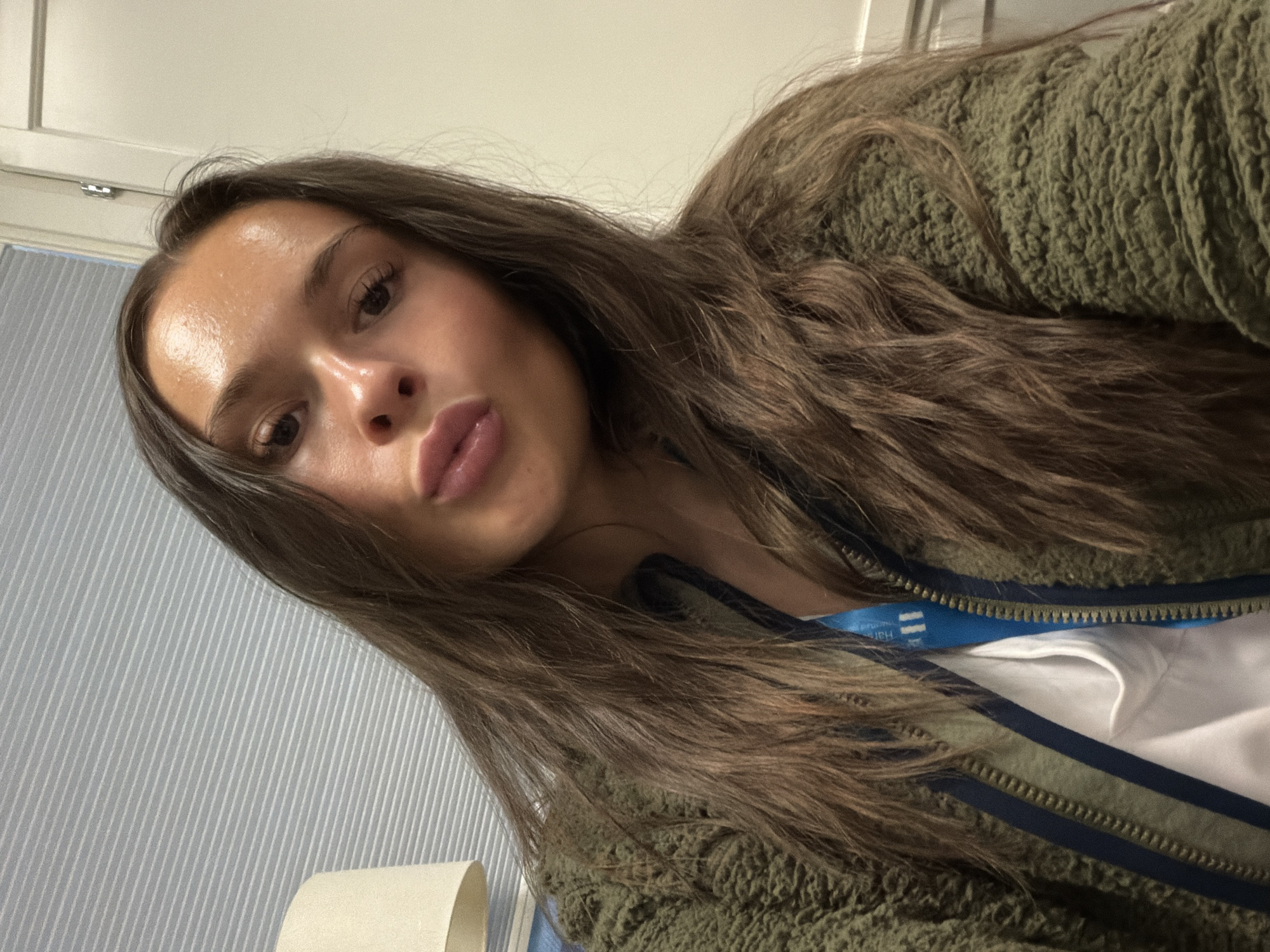 A young woman with long brown hair, wearing a fuzzy green jacket over a white shirt and a blue lanyard, puckers her lips for a selfie. The background appears to be an indoor setting, with horizontal blinds on the left and a white door or wall on the right. The lighting suggests an interior space, possibly an office or a room in a building. There is no indication of the specific location in Harstad, Norway.