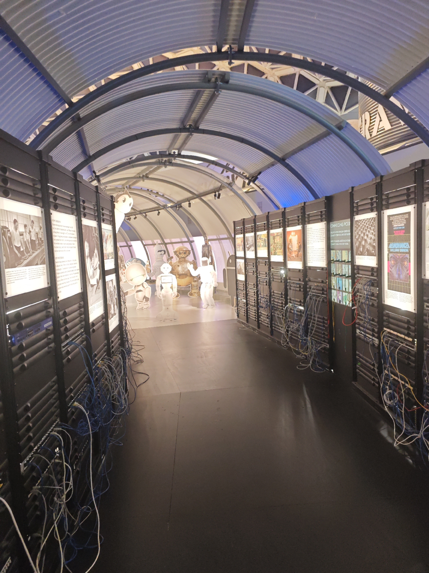 The image depicts a hallway with a corrugated metal ceiling and walls lined with server racks. Informational posters are displayed on the racks, and various robots are visible in the background, including one resembling Astro Boy, another that appears to be Bender from Futurama, and several humanoid white robots. Blue and white network cables are connected to the server racks and spill onto the floor. The scene suggests an exhibition or display related to robotics or technology. This hallway is located within the Philips Museum in Eindhoven, Netherlands, as indicated by the "Connecting Pictures" exhibit visible on one of the posters, a reference to Philips' early innovations in imaging technology and its historical ties to the city.