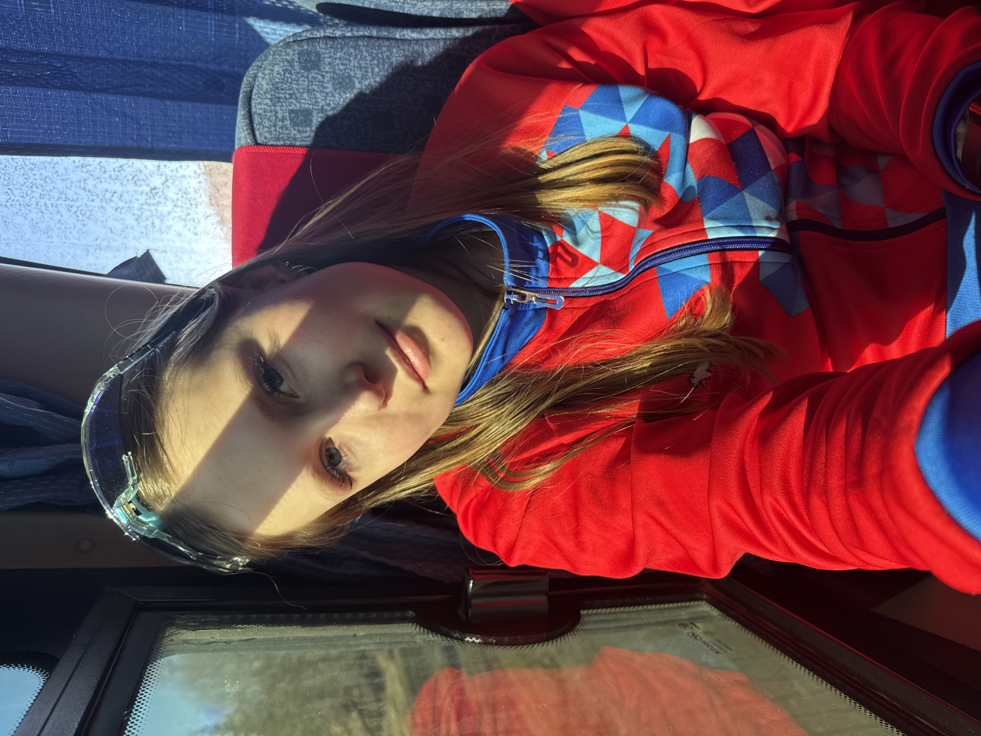 A young fair-skinned girl with light brown hair, wearing clear goggles on her forehead and a red and blue zip-up hoodie, lies at an angle. A bright light casts a shadow across her face. She appears to be inside a vehicle; a bus seat is visible in the background, and a partially visible window is located at the bottom of the frame. It is not possible to confirm her location is Челва, Russia based on visual information.