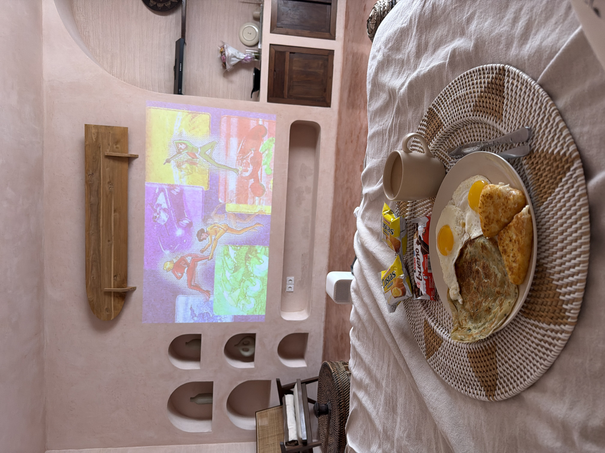 A still life features a bed with a breakfast tray set on it. The tray holds a plate with two fried eggs, hash browns, and a crepe. There's a tan-colored mug and assorted snack packages nearby. The scene is oriented to reveal a section of a wall, where a digital projector casts an image of Renaissance art. A simple wooden shelf adorns the wall to the left of the projected image. Below the projection, semi-circular cubbies are carved into the wall. The wall is tinted in a light, muted pink. To the right of the wall, fragments of furniture and accessories are visible, including dark wooden cabinets and a wicker basket, suggestive of the room's interior design. This scene depicts the interior of a building located in North Kuta, Indonesia.