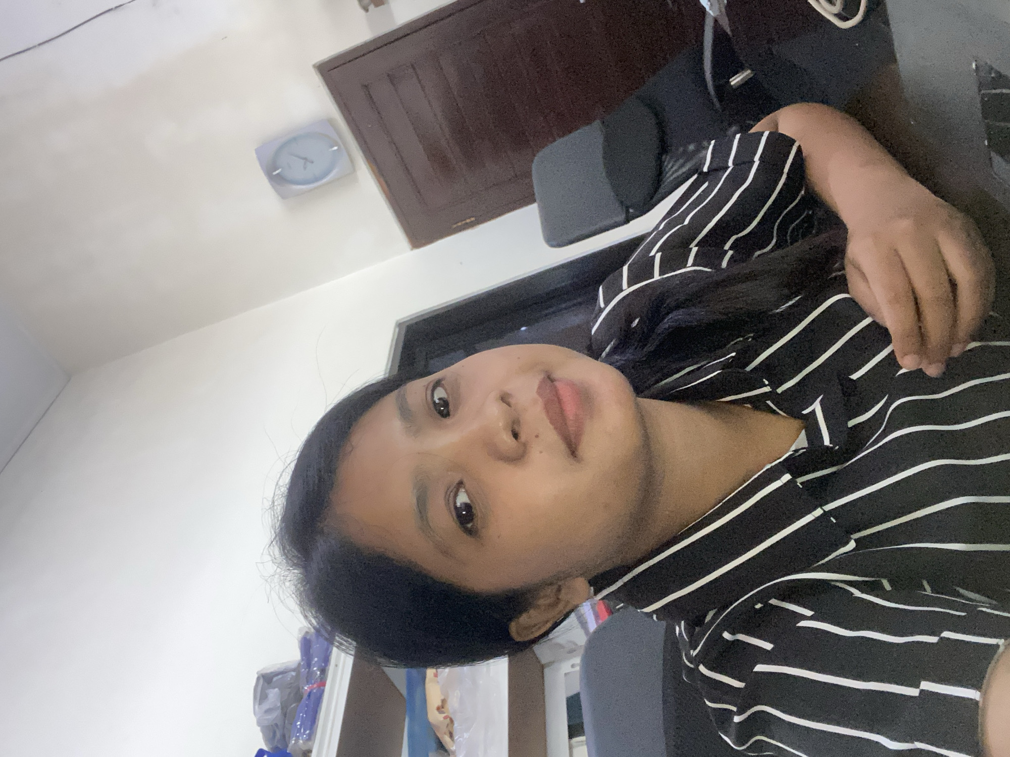 A woman with dark hair is photographed indoors. She is wearing a black and white striped shirt. Behind her, there is a door and a clock on the wall. The location is determined to be Negara, Indonesia.