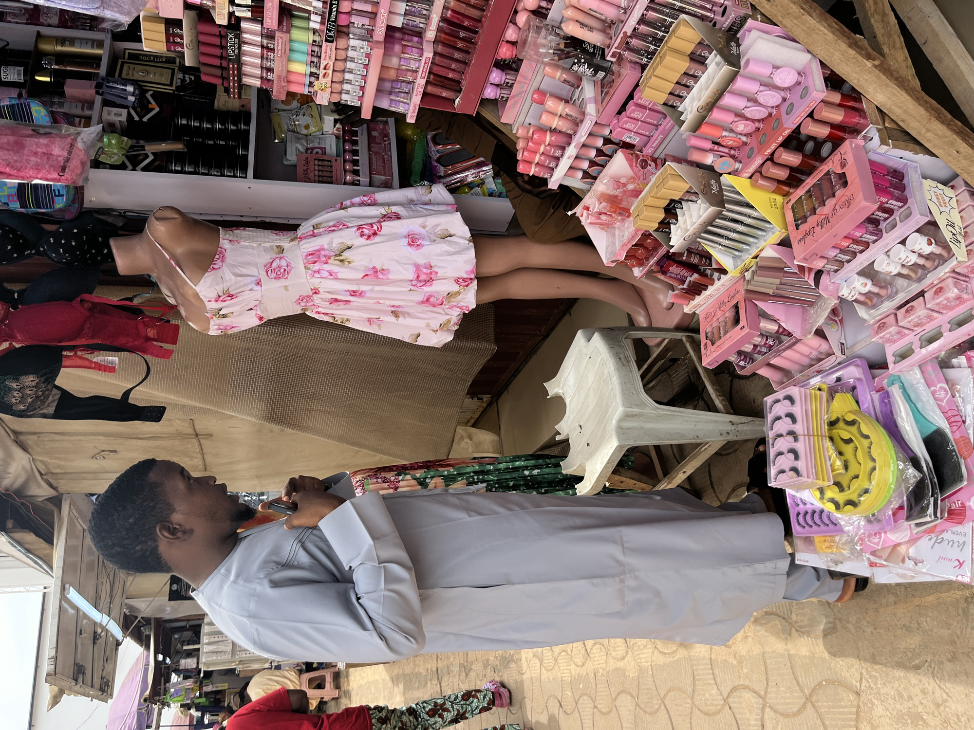This image depicts a market stall in Abuja, Nigeria, specializing in cosmetics and clothing. In the foreground, a man wearing a light grey traditional outfit, possibly a "babban riga," is viewed from the side, looking down at his mobile phone. He has a beard and short hair. To his right, a mannequin displays a pink floral sundress. Behind the mannequin, various cosmetic products, predominantly lipsticks and lip glosses in pink and nude shades, are neatly arranged on shelves and stands. Several bras in red and black are also visible hanging above the cosmetics. The market appears to be outdoors or semi-outdoors, with natural lighting. In the background, other market stalls and people can be seen, suggesting a bustling commercial environment. The ground is paved with interlocking bricks.