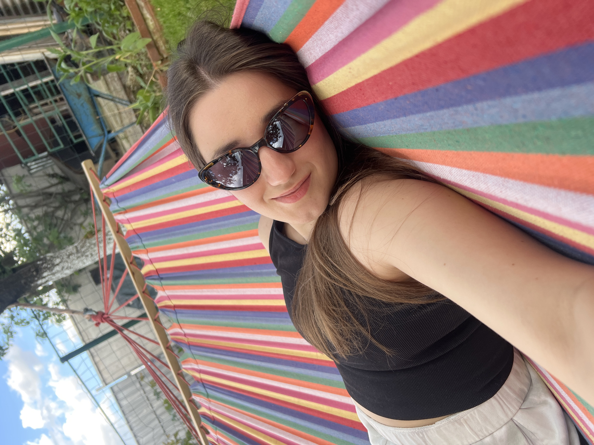 A young woman is lying in a colorful striped hammock outdoors. She is wearing sunglasses, a black tank top, and white pants. The hammock is suspended from a wooden frame with red ropes. The background includes a building with windows, greenery, and the sky. The photo appears to have been taken from the woman's perspective, looking upwards. The location is possibly Busmantsi, Bulgaria, based on the prompt, but there is no visual confirmation of this from the image itself.