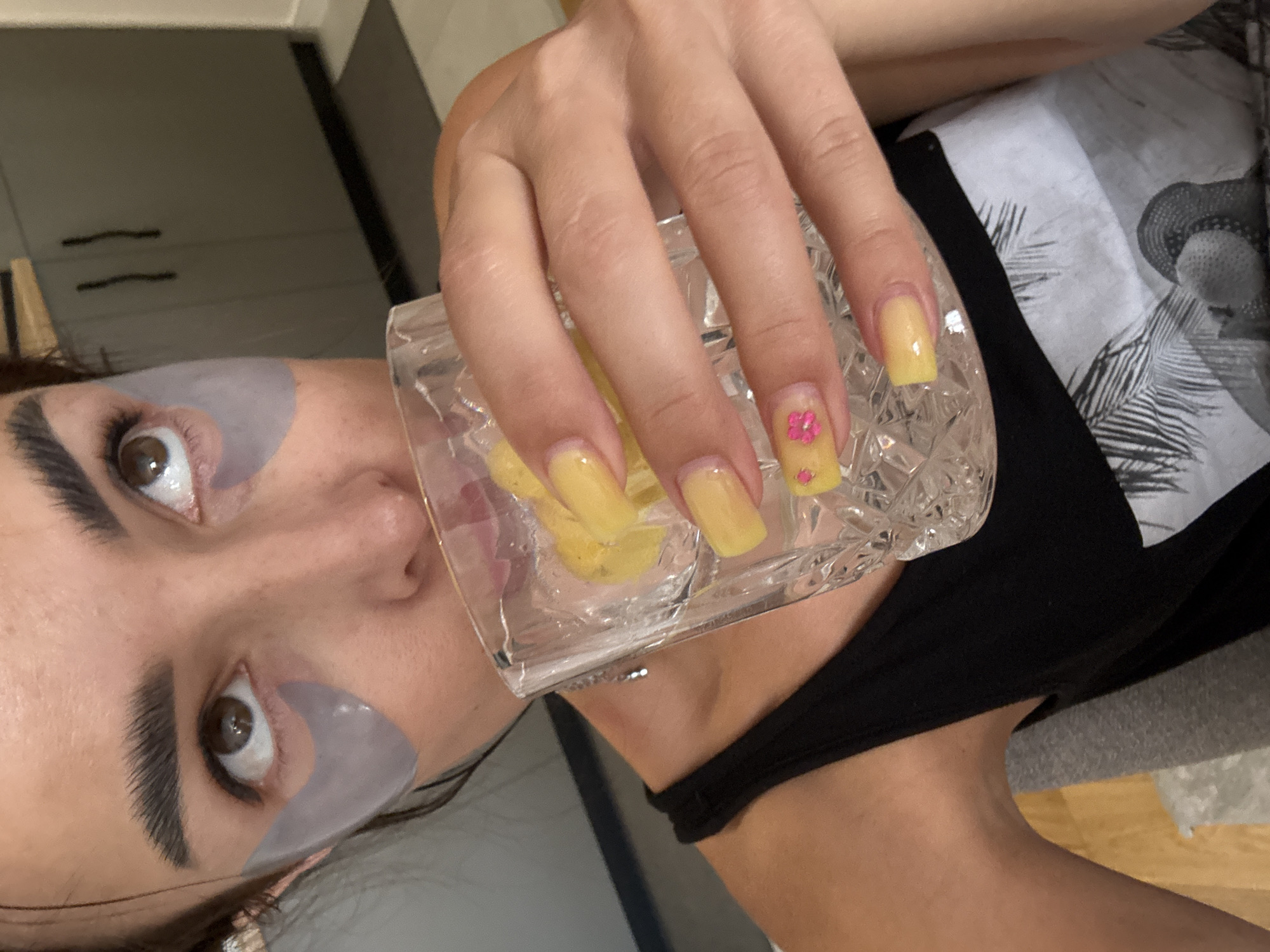 A close-up shot captures a woman in a black tank top as she drinks from a glass of ice water with lemon slices. She is wearing under-eye patches, and her fingernails are painted a bright yellow with small pink flower accents. The background includes kitchen cabinets and a doorway. The image is taken from a high angle, looking down at the woman.