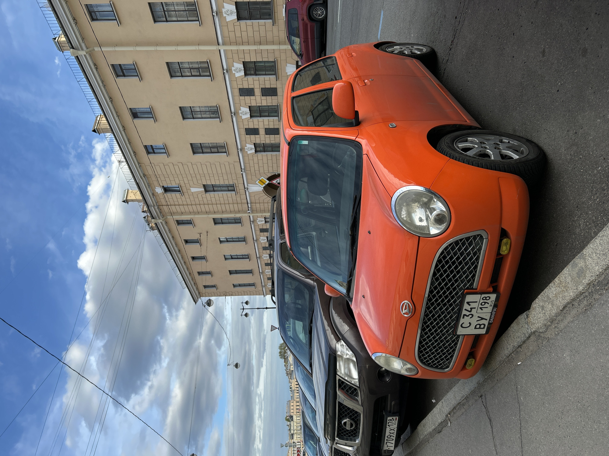 The image shows a street scene in Saint Petersburg, Russia. An orange Daihatsu car is parked on the right side of the frame, partially obscuring a dark SUV behind it. The license plate of the orange car reads "C 341 BY 198 RUS". Behind the vehicles, a light-colored building with multiple windows extends upwards. Above the building, a cloudy sky is visible, with electrical wires running across the scene. The image is taken at an angle, tilting the perspective to the right.