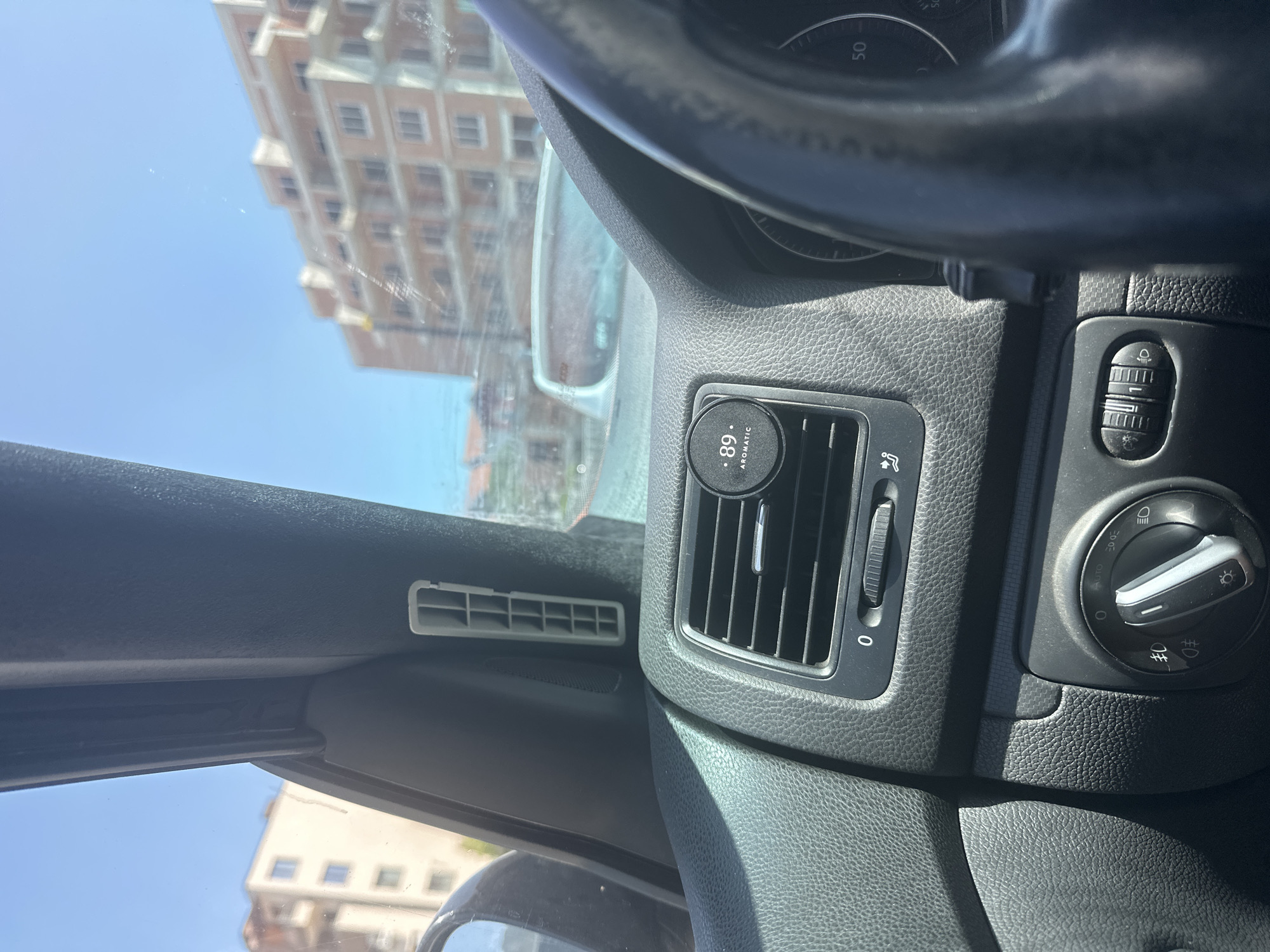 The image shows the interior of a car, looking out through the windshield. In the foreground, the dashboard is visible, with a car air vent on the right side of the image. A circular black air freshener with "89 AROMATIC" printed on it is attached to the air vent. The steering wheel is partially visible on the right. In the background, there are buildings under construction, a clear blue sky, and some trees. The scene is captured during the daytime. The presence of construction suggests it may be a developing area within the city.
The specific location in Vratsa, Bulgaria cannot be determined from the image alone.
