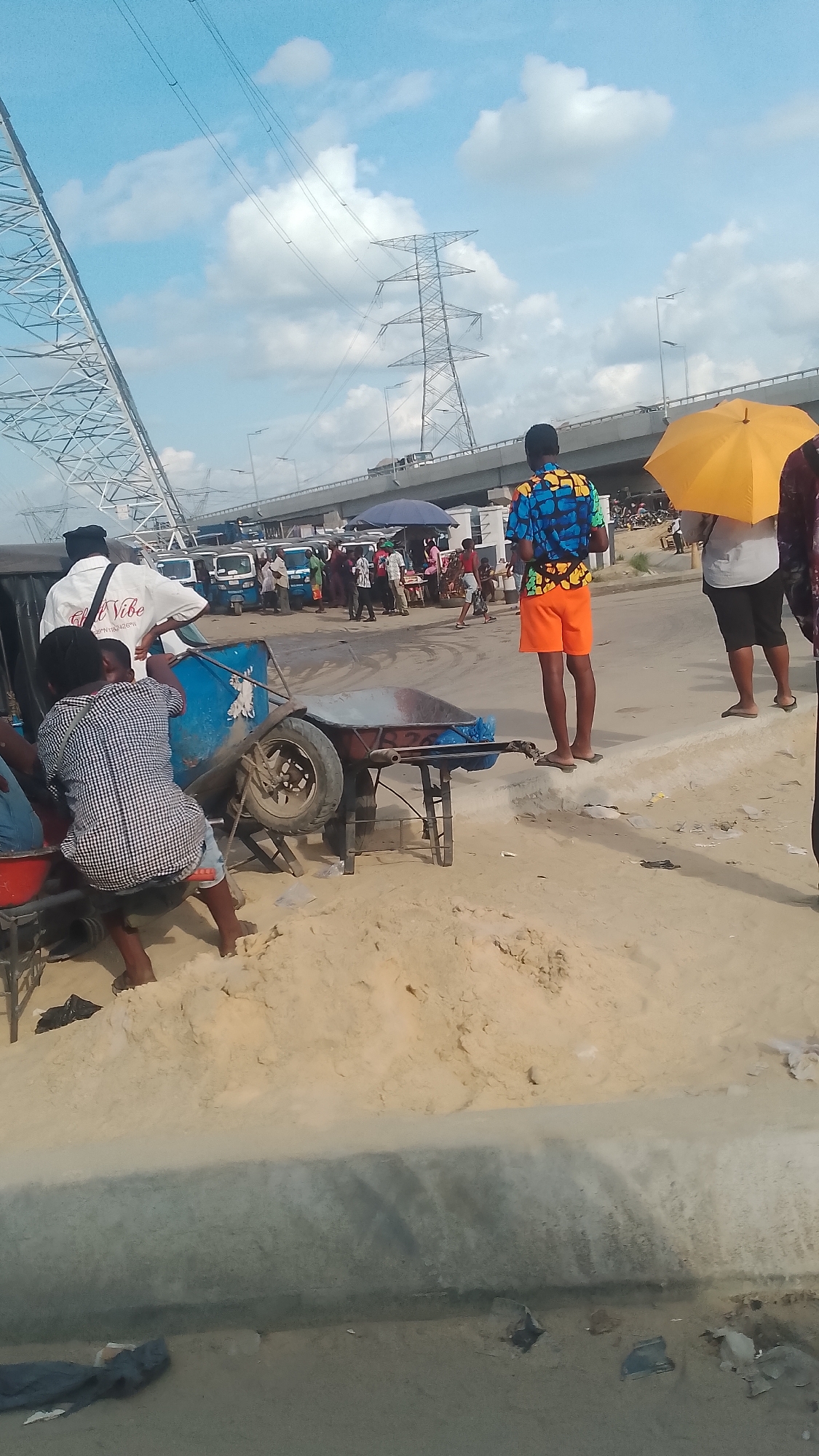 The image captures a street scene in Effurun, Nigeria, under a partly cloudy sky with visible power lines and a bridge in the background. Several people are present; one individual stands out in bright orange shorts and a colorful shirt. Another person is holding a yellow umbrella. In the foreground, there's a makeshift cart or vehicle with people sitting and interacting around it. The ground is covered with sand and debris. There are also a few auto rickshaws in the vicinity.