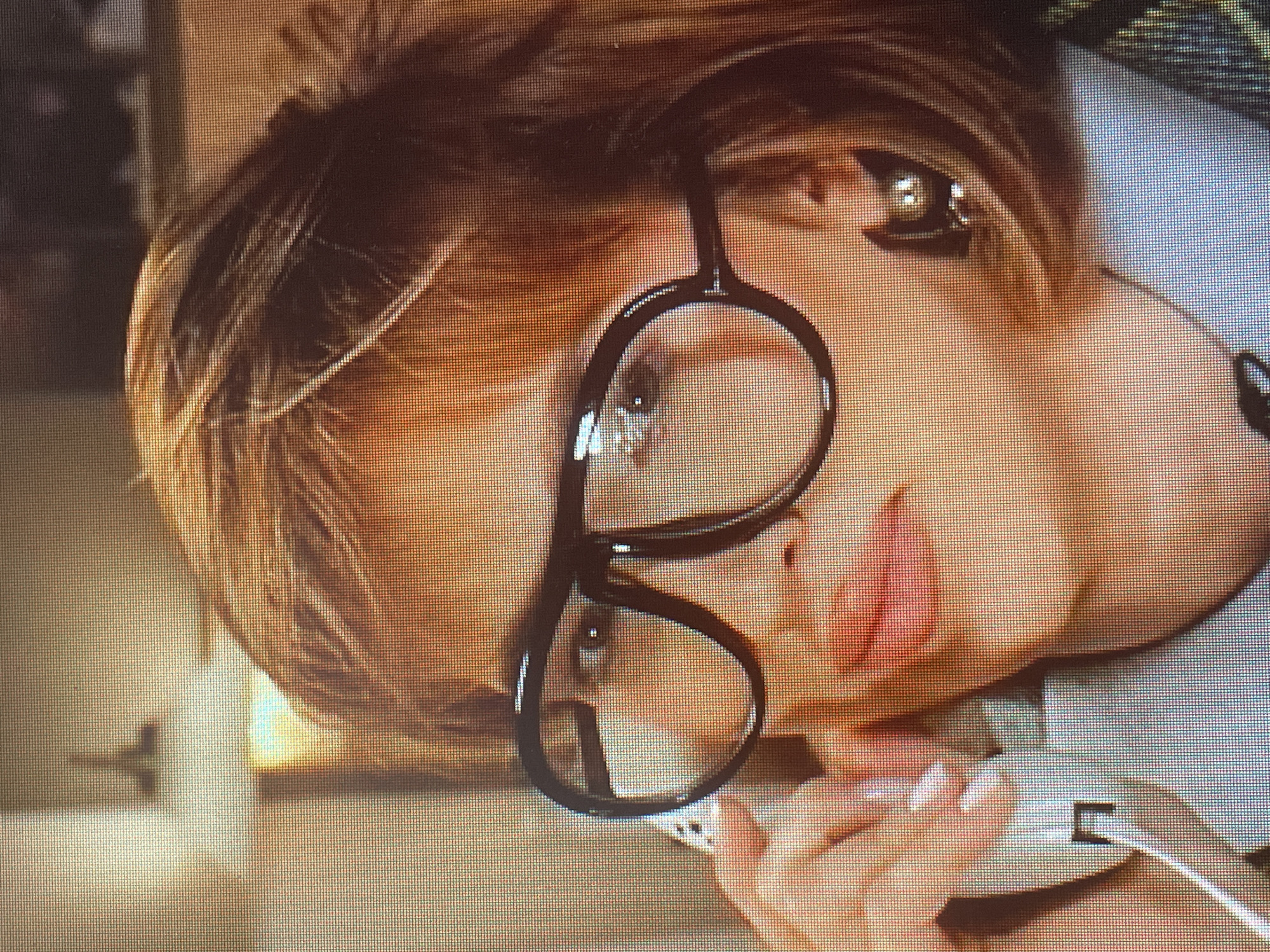 The media file shows a close-up, angled shot of a woman wearing large, black, circular-framed glasses. Her hair, appearing to be a light brown or blonde, is styled in a way that obscures part of her face. She is holding a white phone to her ear with her right hand, and her left hand is also visible, resting near the phone. The woman's lips are slightly parted and appear to be painted with a pink or coral lipstick. Her eyes are visible through the lenses of her glasses, and one eye has a subtle reflection in the pupil.
The background is blurred but suggests an indoor setting with warm lighting, possibly artificial. There are hints of muted colors and soft shapes, making it difficult to discern specific details of the environment. A faint shadow of what might be a coat rack or some other object is visible on the left side of the frame.
The overall impression is one of intimacy and perhaps a moment of casual conversation or reflection. The lighting and the close framing create a sense of personal space. There is no visible text in the media file. The context provided suggests the location is Sinyavino, Russia, but there are no visual cues within the image to confirm or deny this. The time of day and weather are not discernible.