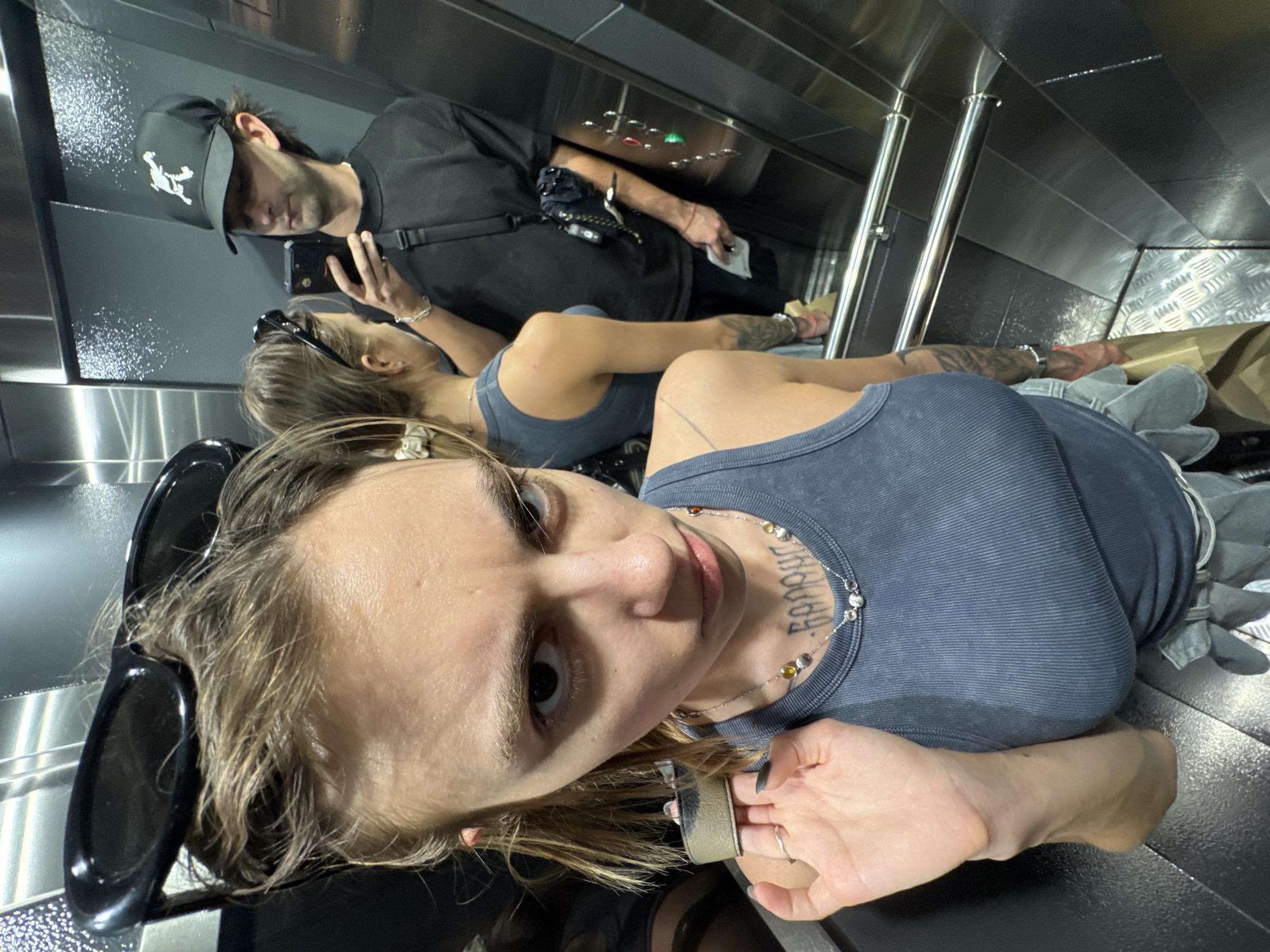 The image depicts three individuals inside an elevator with metallic walls. The foreground is dominated by a young woman with light brown hair, wearing a dark gray tank top, a necklace, and sunglasses perched on her head. Her gaze is directed toward the camera. Behind her is another woman with a similar top, though partially obscured. Further back, a man in a black t-shirt and baseball cap is looking at his mobile phone. The elevator's interior is shiny and stainless steel, with control panels and handrails visible. The city of Moscow, Russia, is not evident in the image.