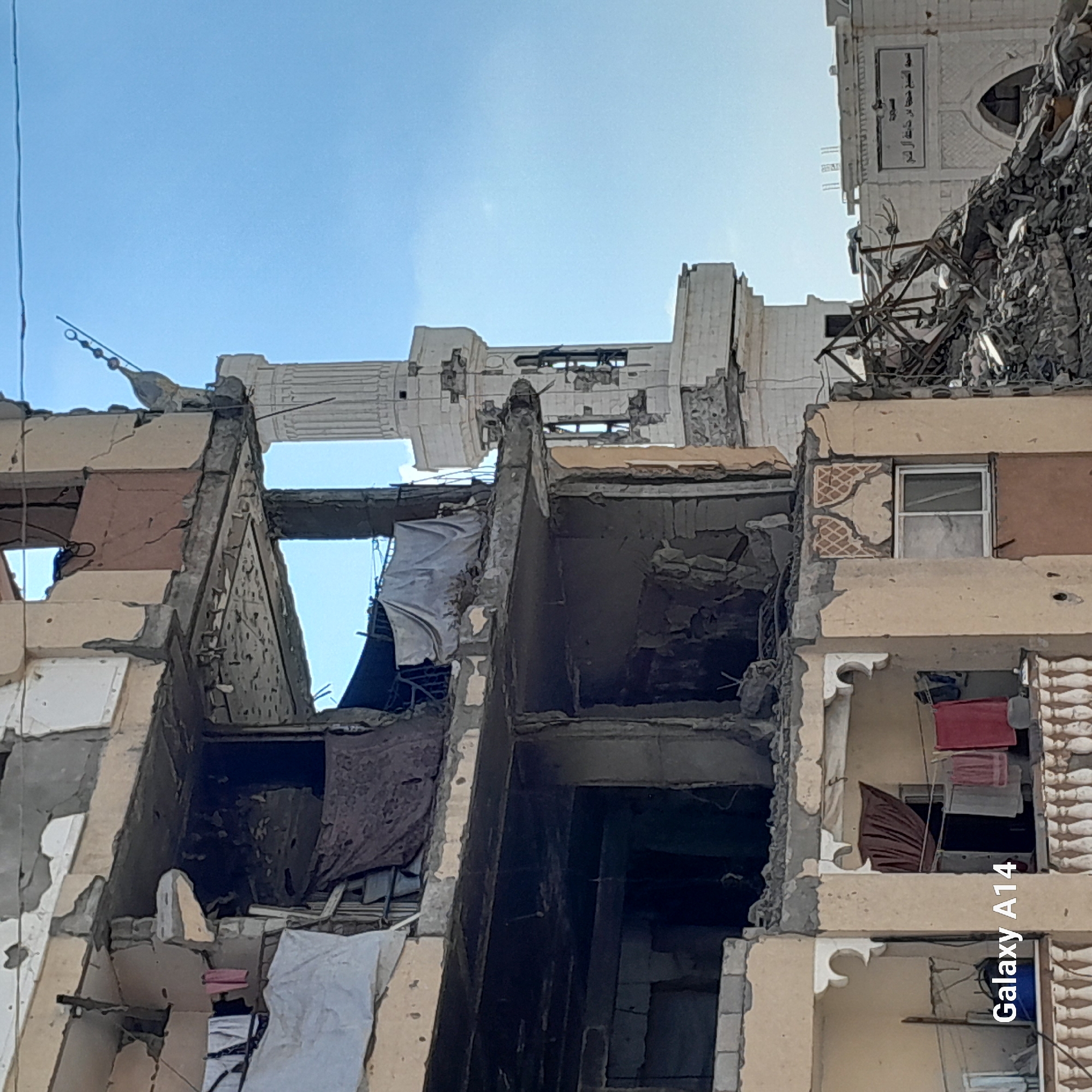 The image captures a scene of destruction in the city of Khan Yunis, Palestinian Territories. A partially collapsed multi-story building dominates the foreground, with exposed concrete, twisted metal, and debris indicating significant damage. Laundry, including towels and clothing, is visible hanging from lines on the remaining balconies, suggesting continued life amidst the ruins.
In the background, a minaret of a mosque stands relatively intact, though also showing signs of damage, against a clear blue sky. The mosque appears to be a pale, light-colored structure. To the right of the minaret, a building with Arabic inscriptions is partially visible, also exhibiting damage and debris. The overall impression is one of a war-torn urban environment, with residential structures and a religious building bearing the brunt of conflict. The "Galaxy A14" watermark is present in the lower left corner of the image.