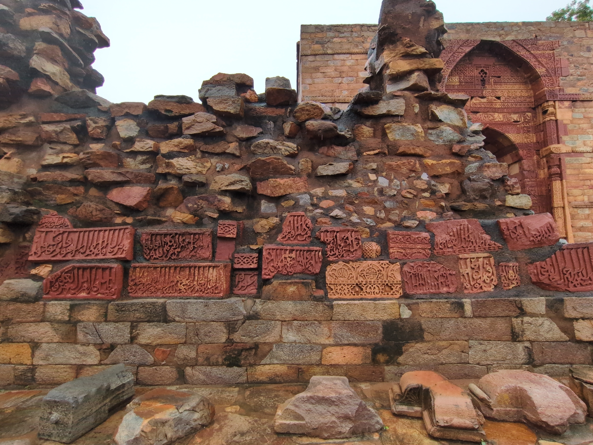 A stone wall with intricate Arabic calligraphy carvings is displayed prominently in the foreground. The carvings are reddish-brown and are set against a backdrop of rough, irregular stones. Below the carved section, the wall transitions to a more uniform brickwork pattern with stones of various earthy tones. In the lower foreground, several large, rough-hewn stones and fragments of what appear to be terracotta or brick lie scattered on the ground. The background reveals a portion of an ancient brick structure with decorative arches and more calligraphy, suggesting a historical site. The sky above is overcast. This scene is located at the Qutub Minar complex in Delhi, India, a UNESCO World Heritage site known for its historical monuments and Islamic architecture.