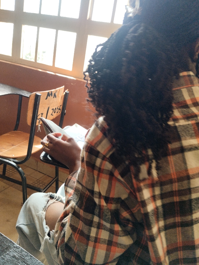 A person sits at a desk near a window. The person is wearing a flannel shirt and ripped jeans, holding a book or notebook. The chair next to them is light brown with a black frame. Inscribed on the chair is the abbreviation "MK" over the date "CT/2055." The window has multiple panes.